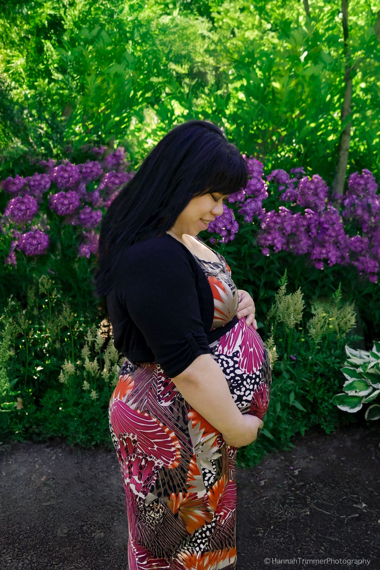 Pregnant woman in floral dress cradles baby bump in garden with purple flowers.