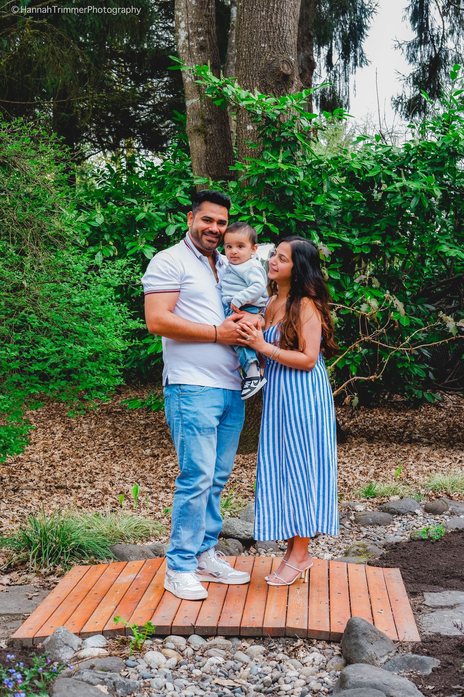 A young family with a baby poses together on a wooden bridge surrounded by lush green garden.
