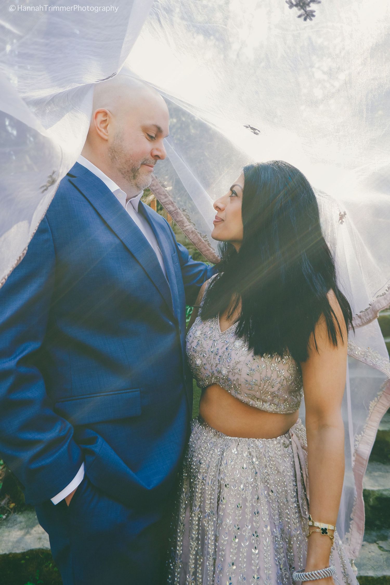 Groom in navy suit and bride in silver lehenga share an intimate moment beneath a sheer flowing veil outdoors.