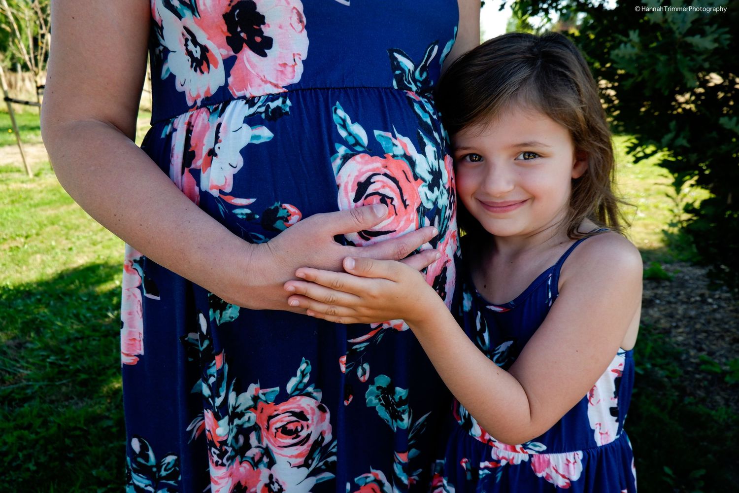 A smiling young girl in a navy floral dress gently holds her pregnant mother's baby bump outside.