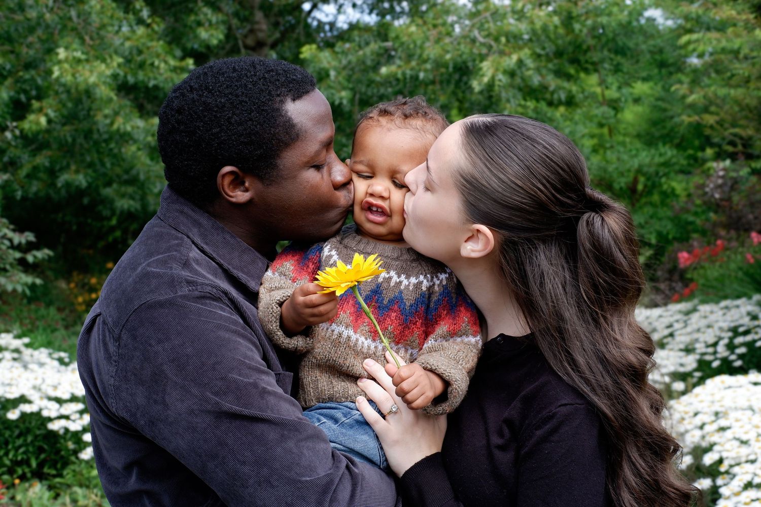 Parents kissing their laughing baby holding a yellow flower in a garden setting.