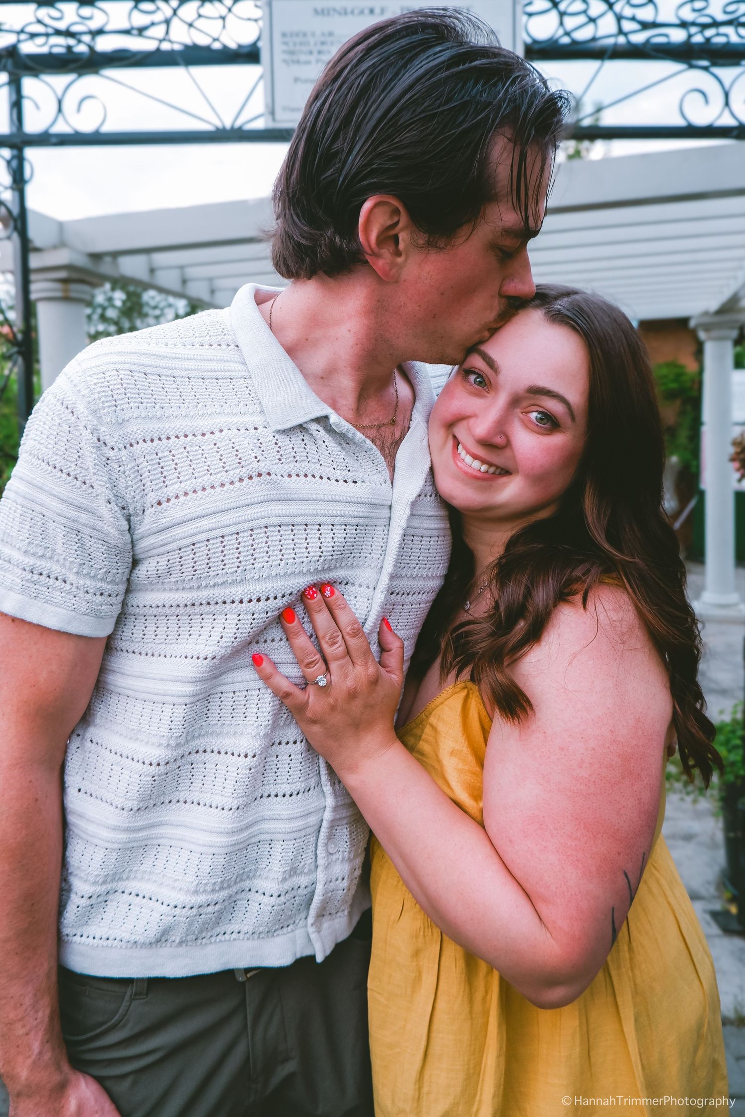 Man kissing smiling woman wearing yellow dress showing engagement ring outdoors.