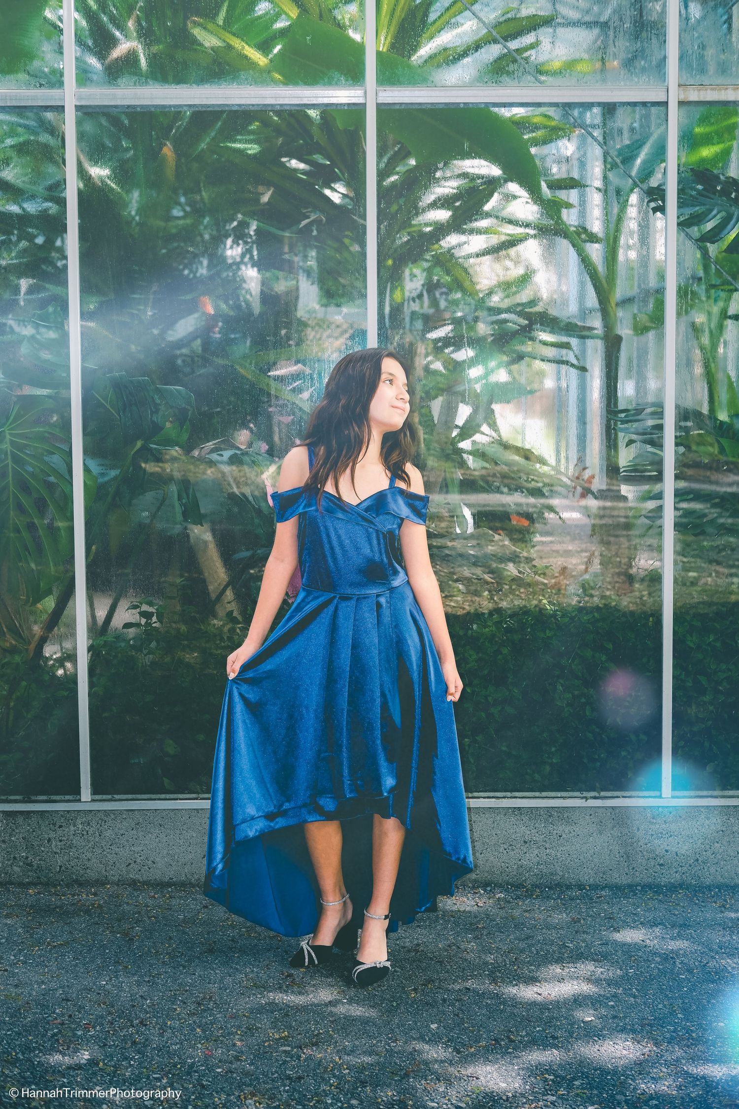 Woman in blue satin off-shoulder high-low dress posing near greenhouse window.