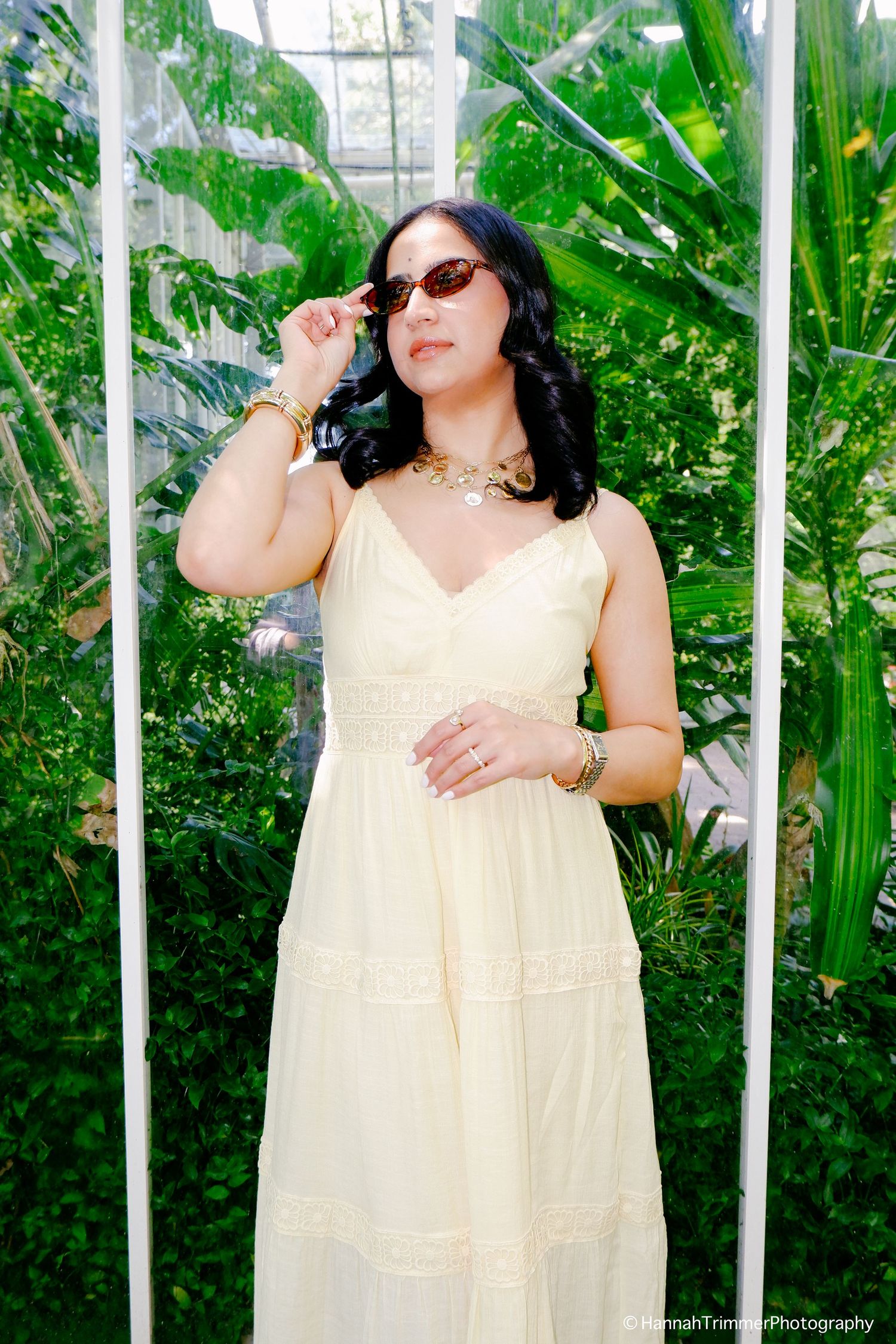 Woman in cream sundress adjusting sunglasses while posing in a bright tropical greenhouse.