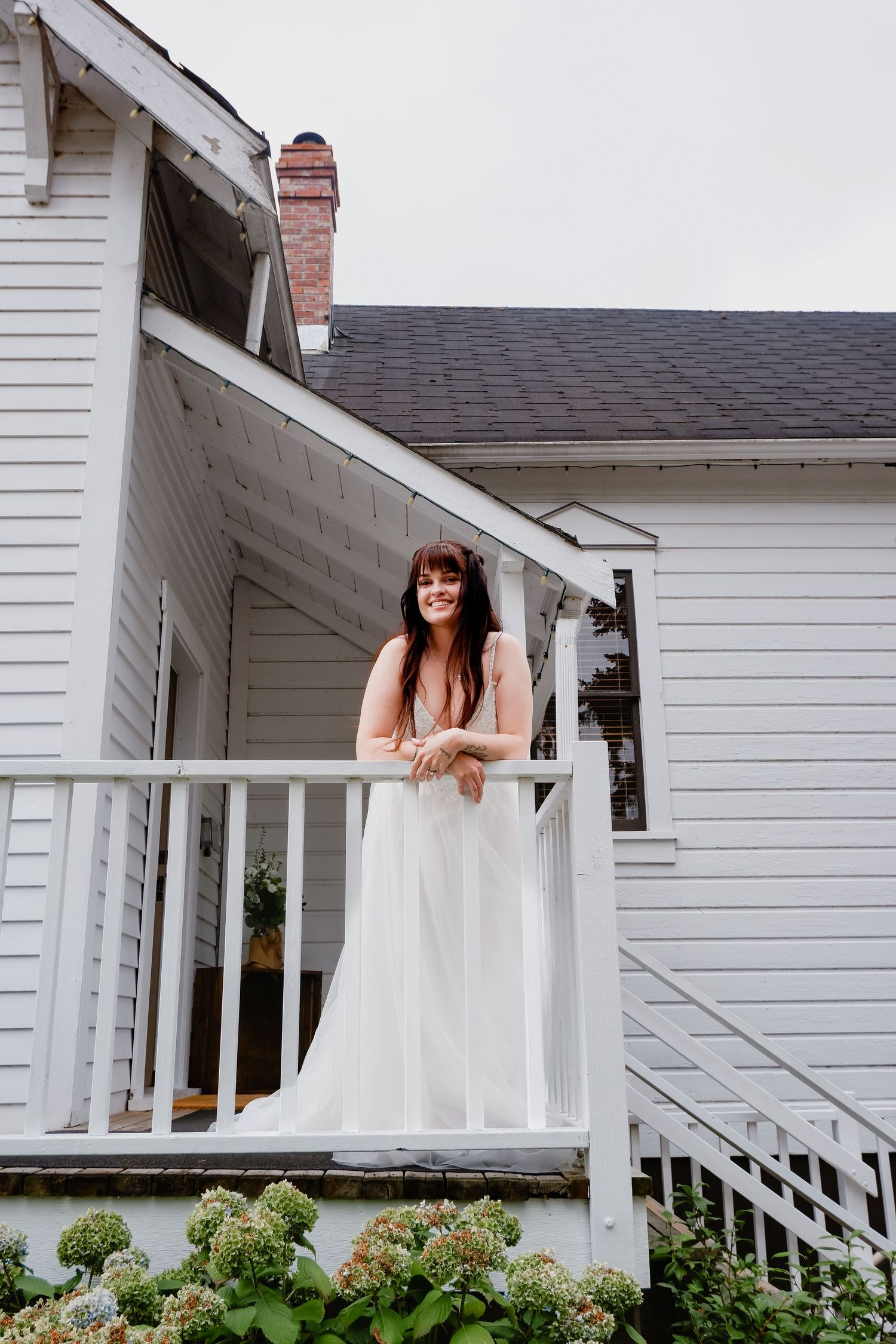 Smiling bride in white gown leaning on porch railing of white farmhouse with hydrangeas below.