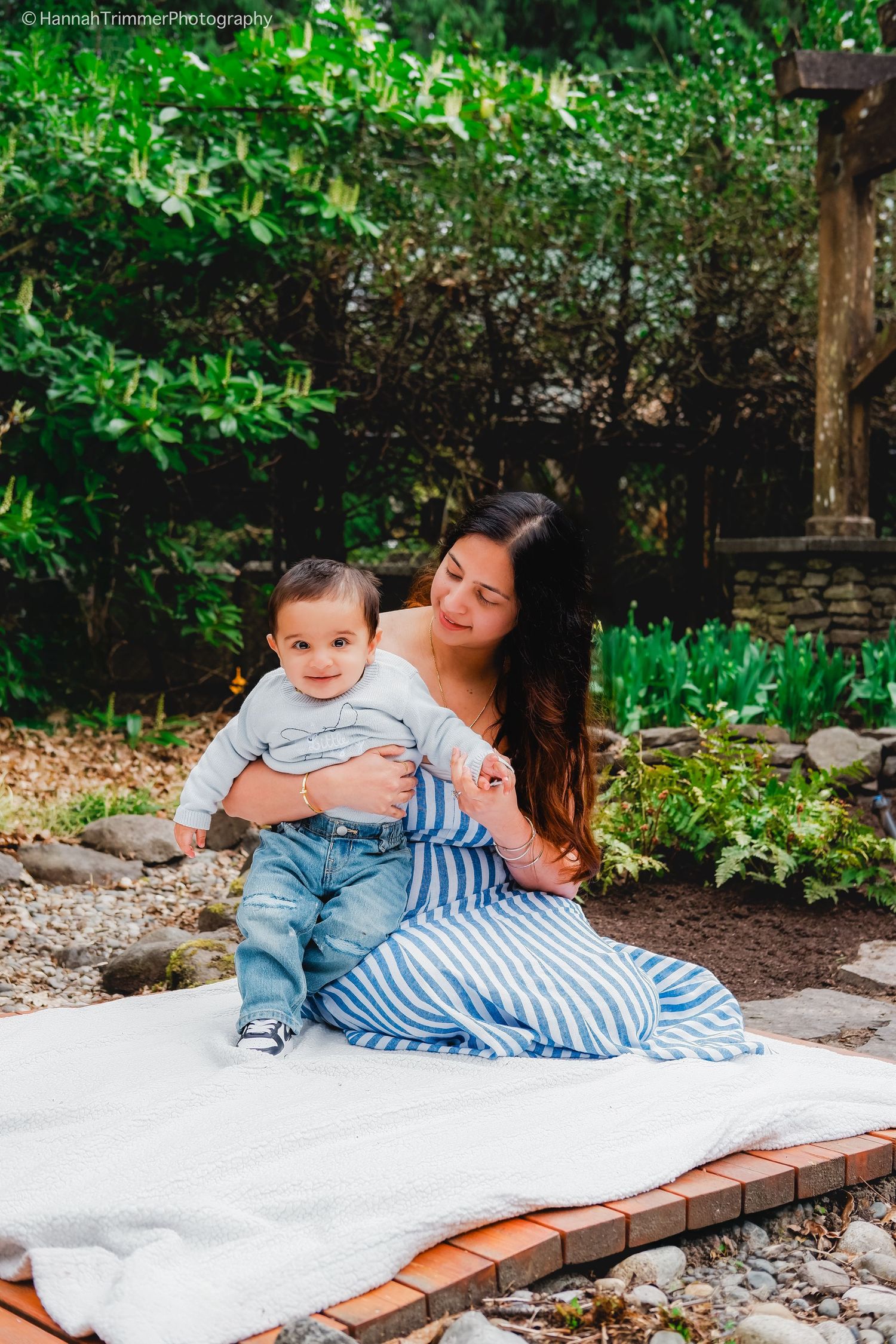 A mother in a blue striped dress sits on a white blanket outdoors, holding her smiling baby boy.