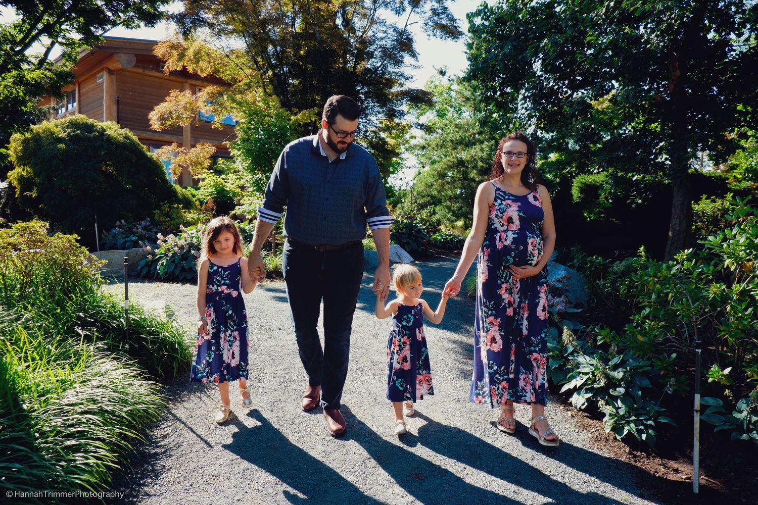 Family of four in matching navy floral outfits walks hand-in-hand along a sunny garden pathway.