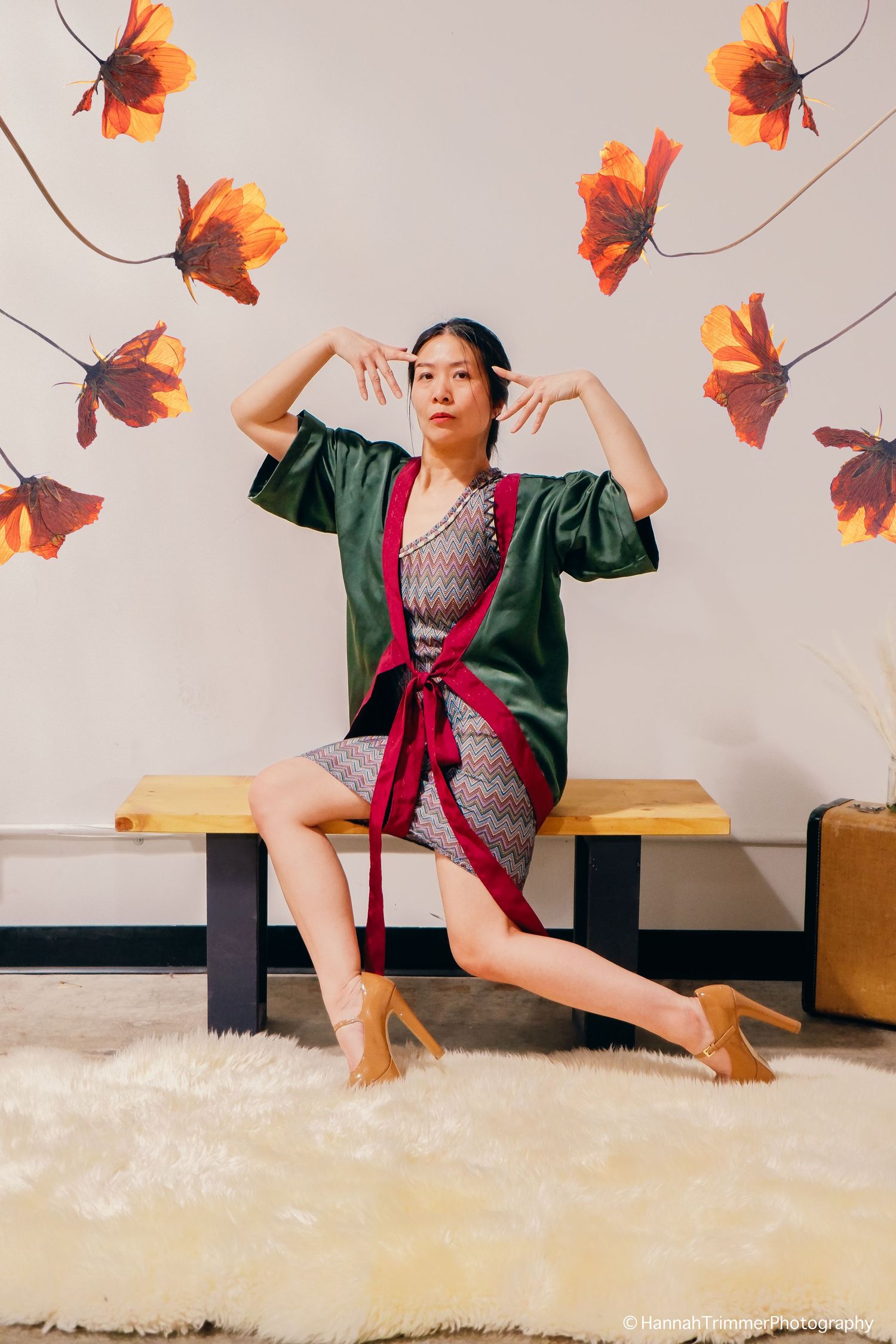 Asian woman in green kimono jacket poses on bench with floral wall art.