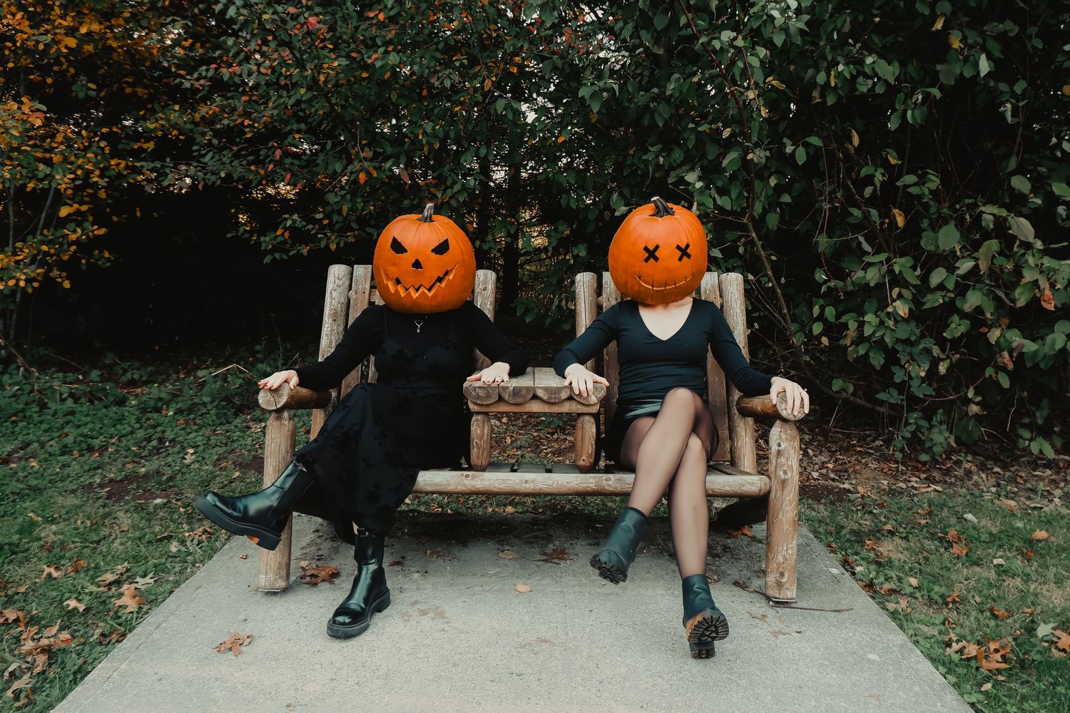 Two people wearing black outfits and carved pumpkin heads sit on a wooden bench outdoors for Halloween.