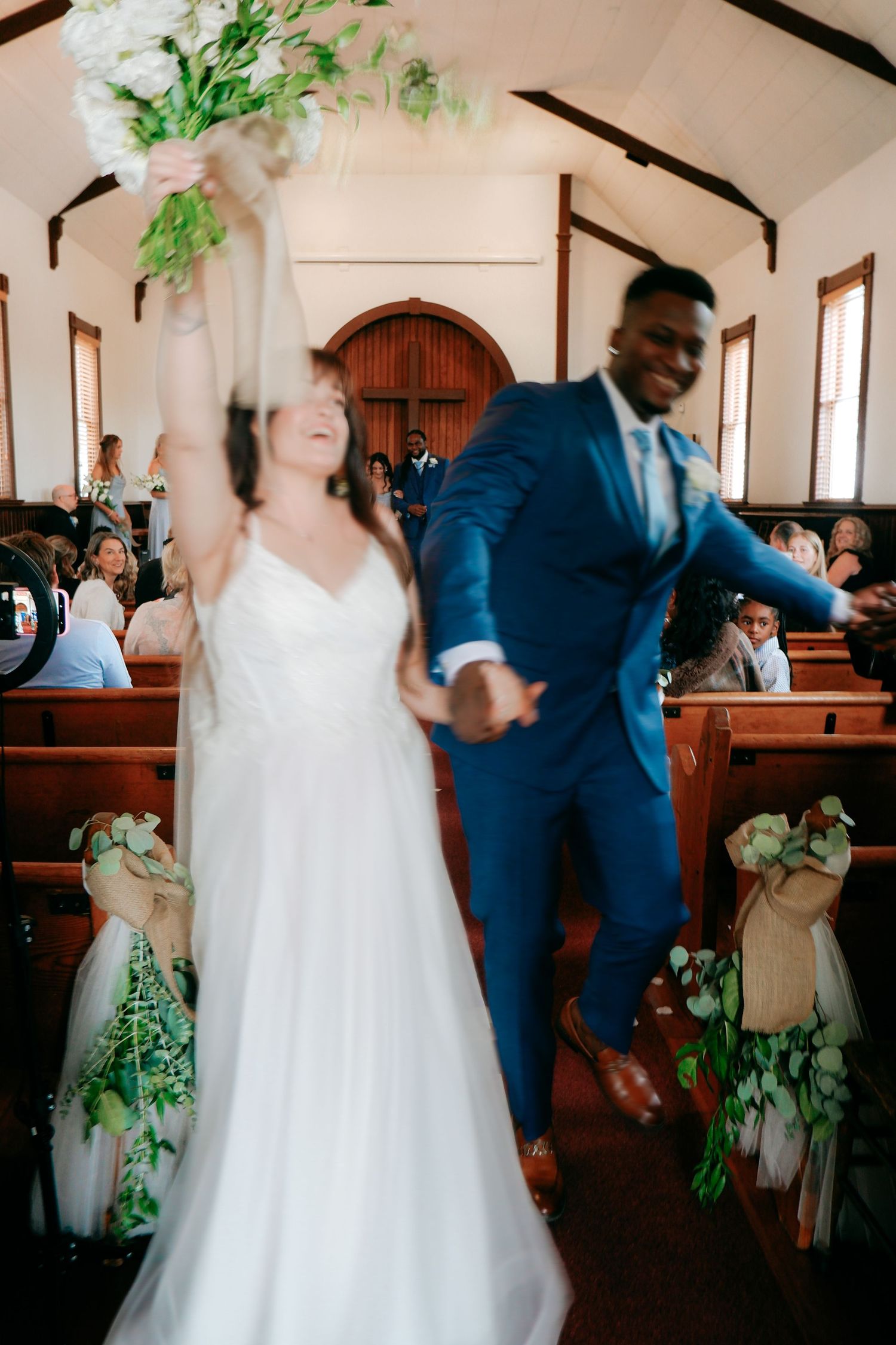 Joyful newlyweds celebrate in church aisle, bride in white gown raises bouquet, groom in blue suit jumps with excitement.