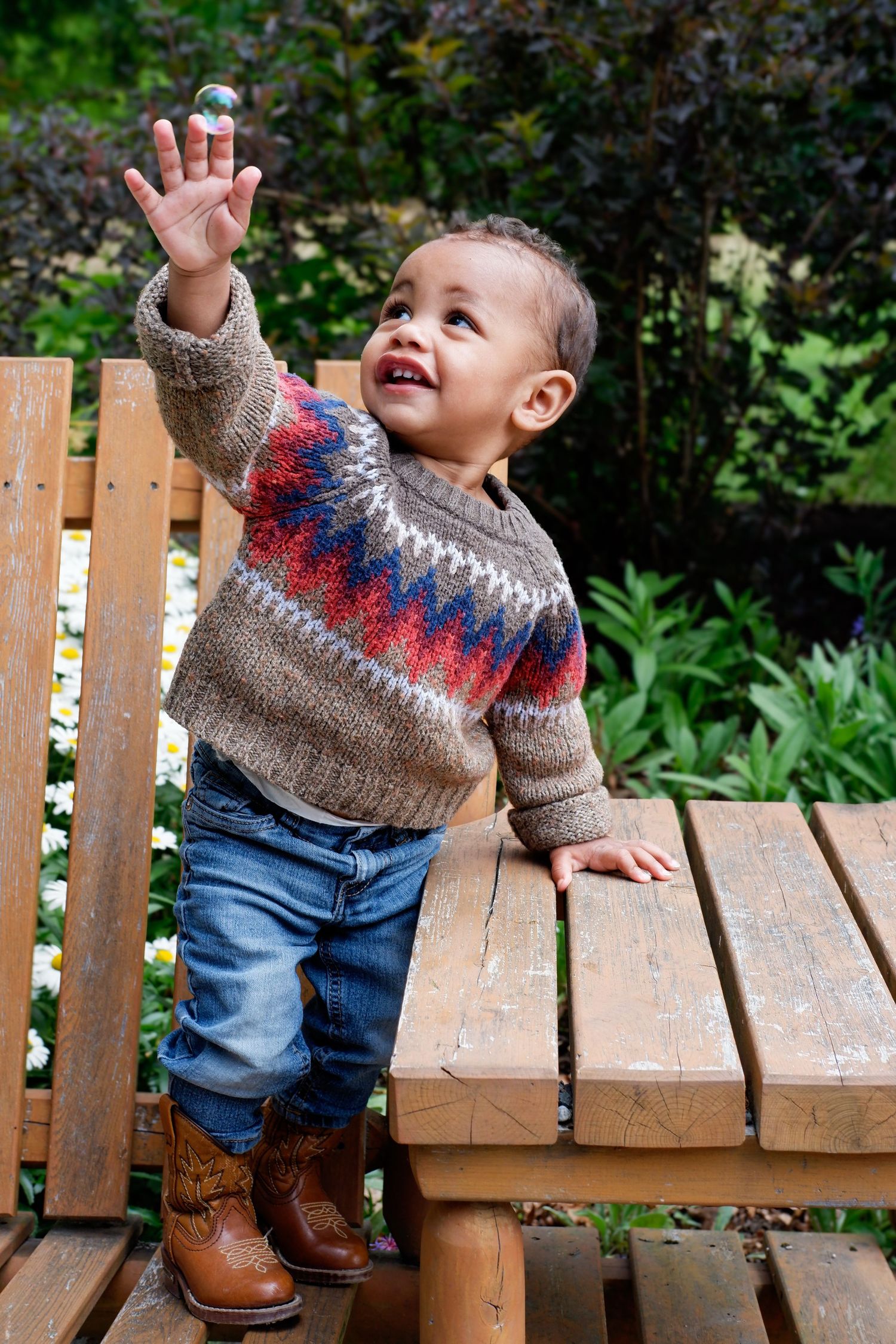 Happy toddler in patterned sweater and cowboy boots reaching up while standing at a wooden bench.