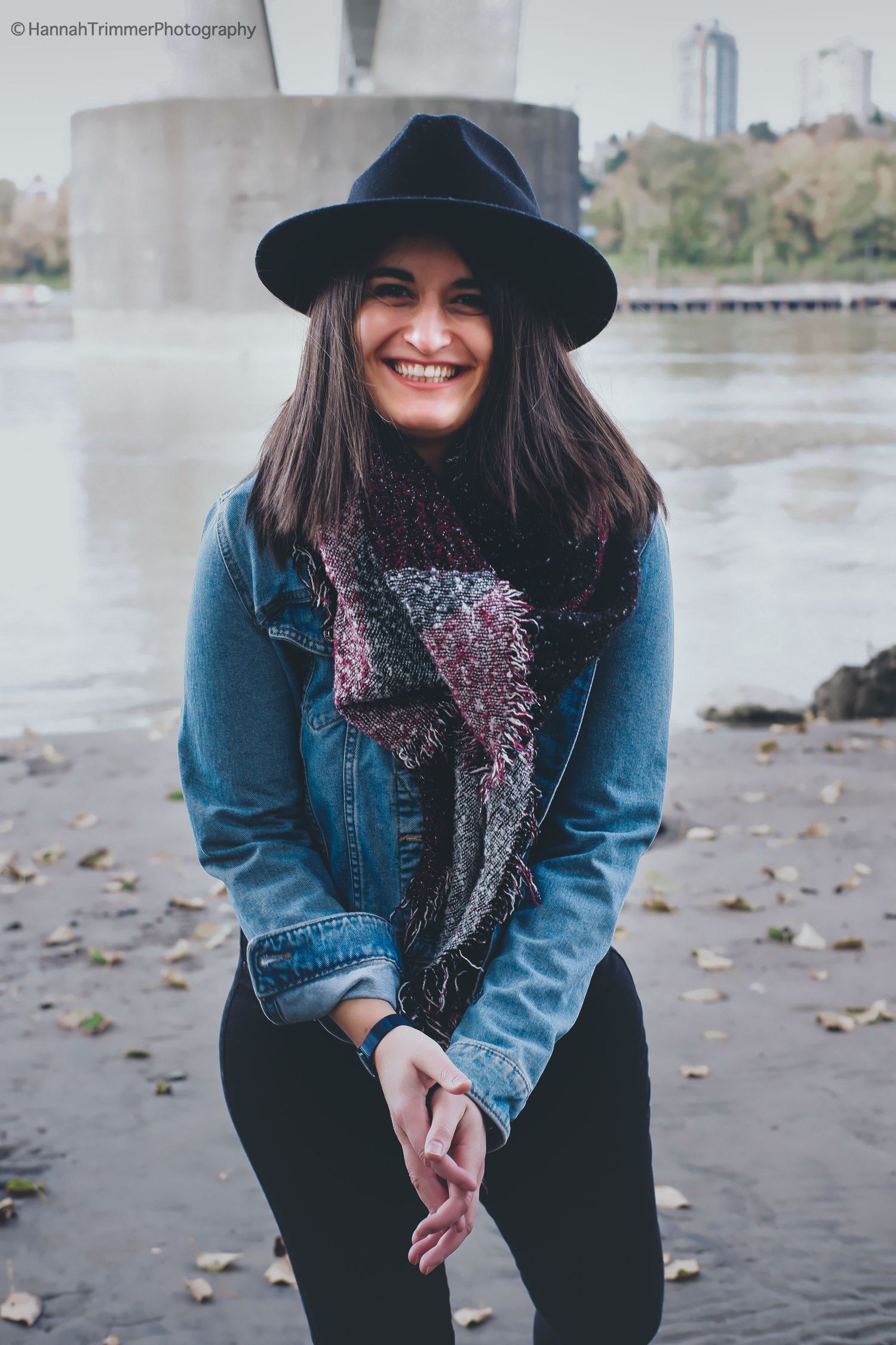 Smiling woman in denim jacket and black hat posing outdoors near a river on an autumn day.