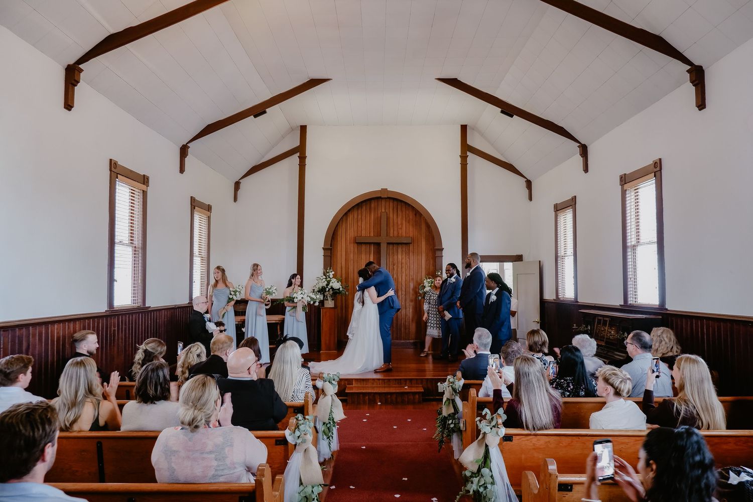 Couple sharing a kiss at the altar in a charming white chapel wedding ceremony with guests seated in wooden pews.