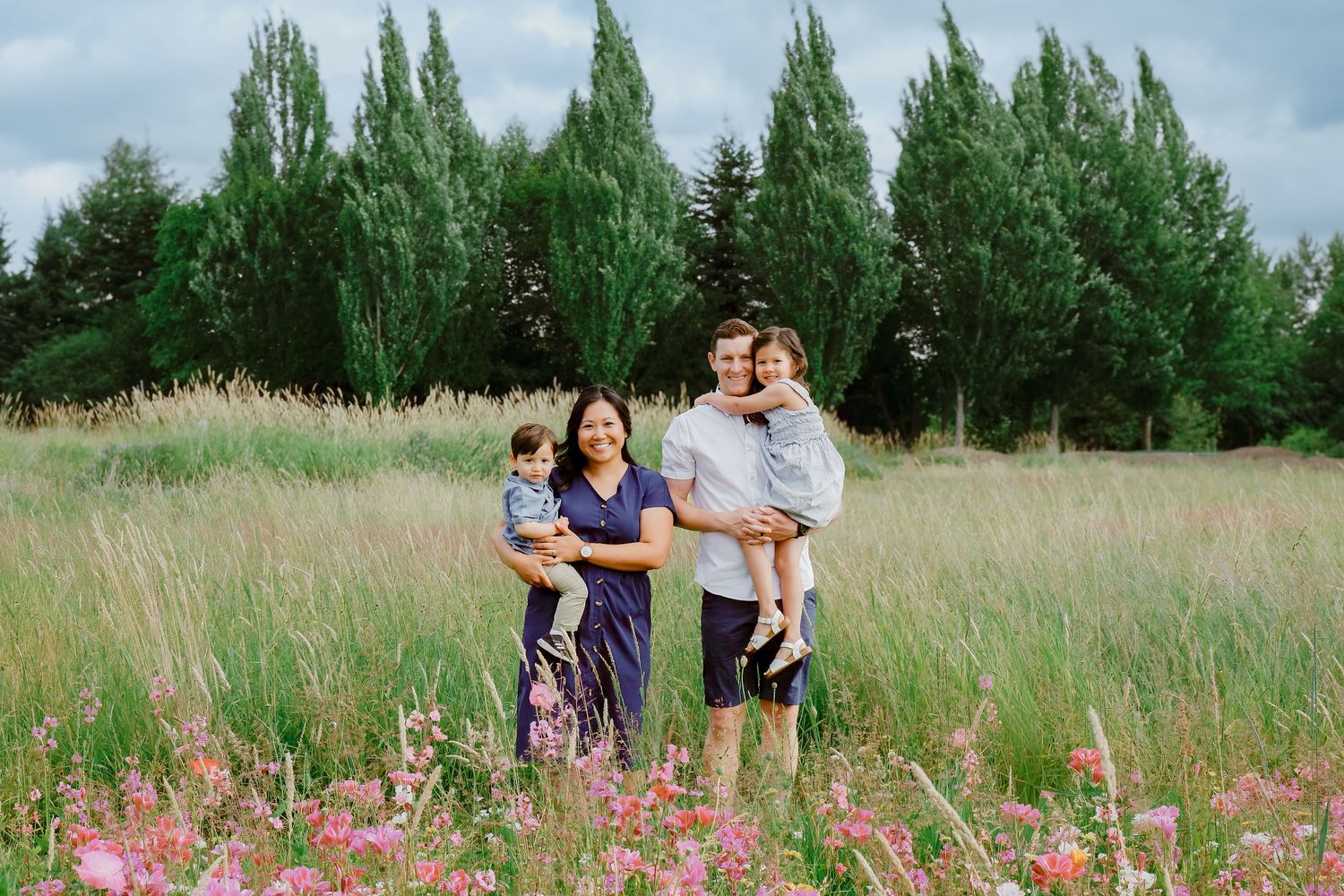 Smiling family of four poses in a lush meadow with pink wildflowers and tall evergreen trees in background.