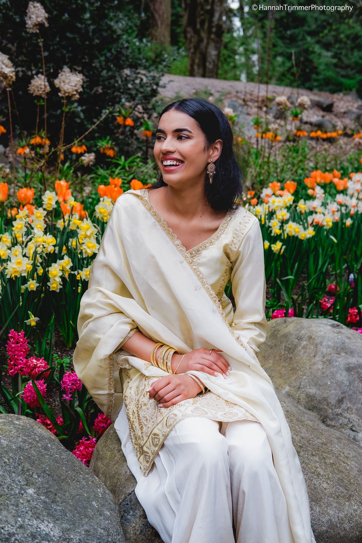 Smiling woman in cream embroidered salwar kameez sits on rock surrounded by colorful spring flowers.