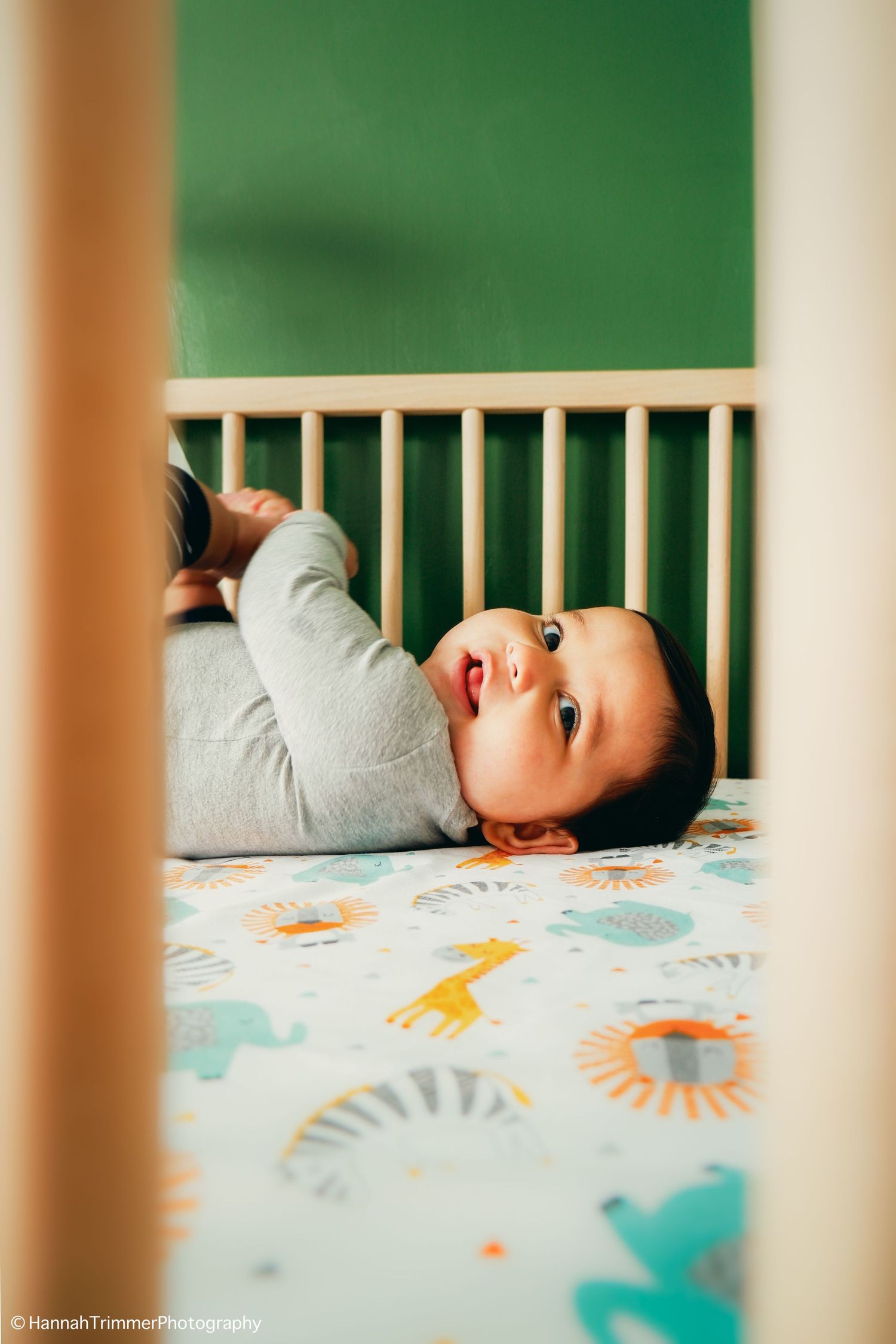 Baby in gray onesie lies in wooden crib with safari-themed bedding.