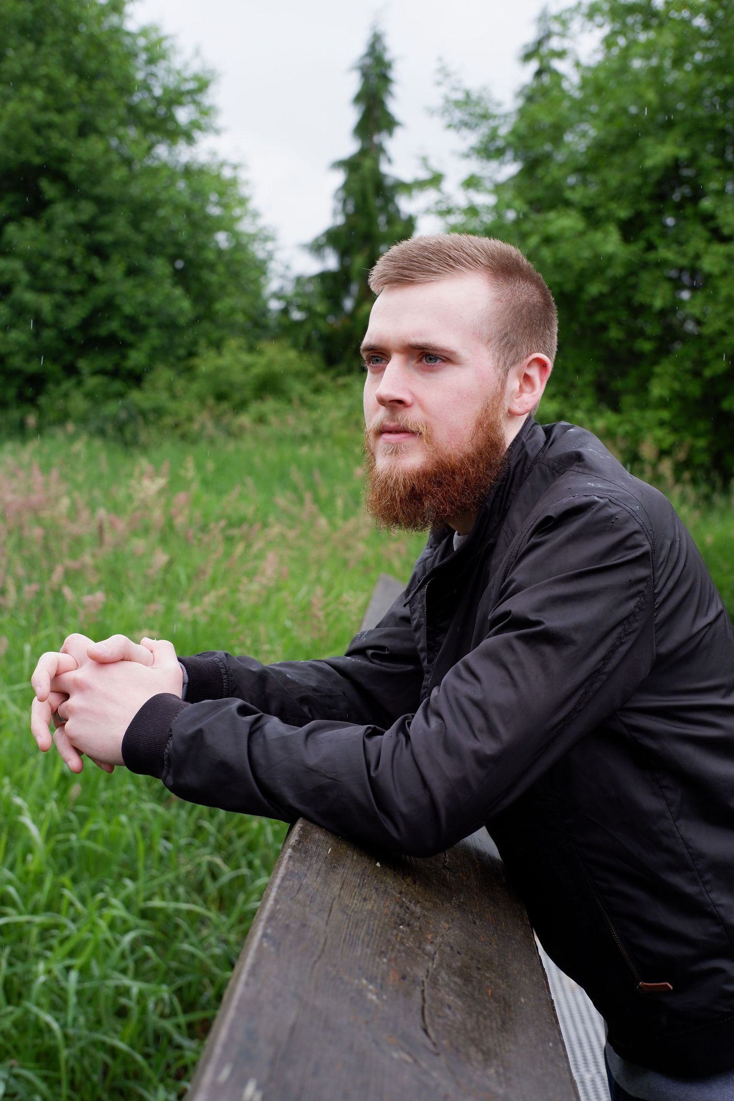 Bearded man in black jacket leaning on wooden fence gazing into green meadow outdoors.