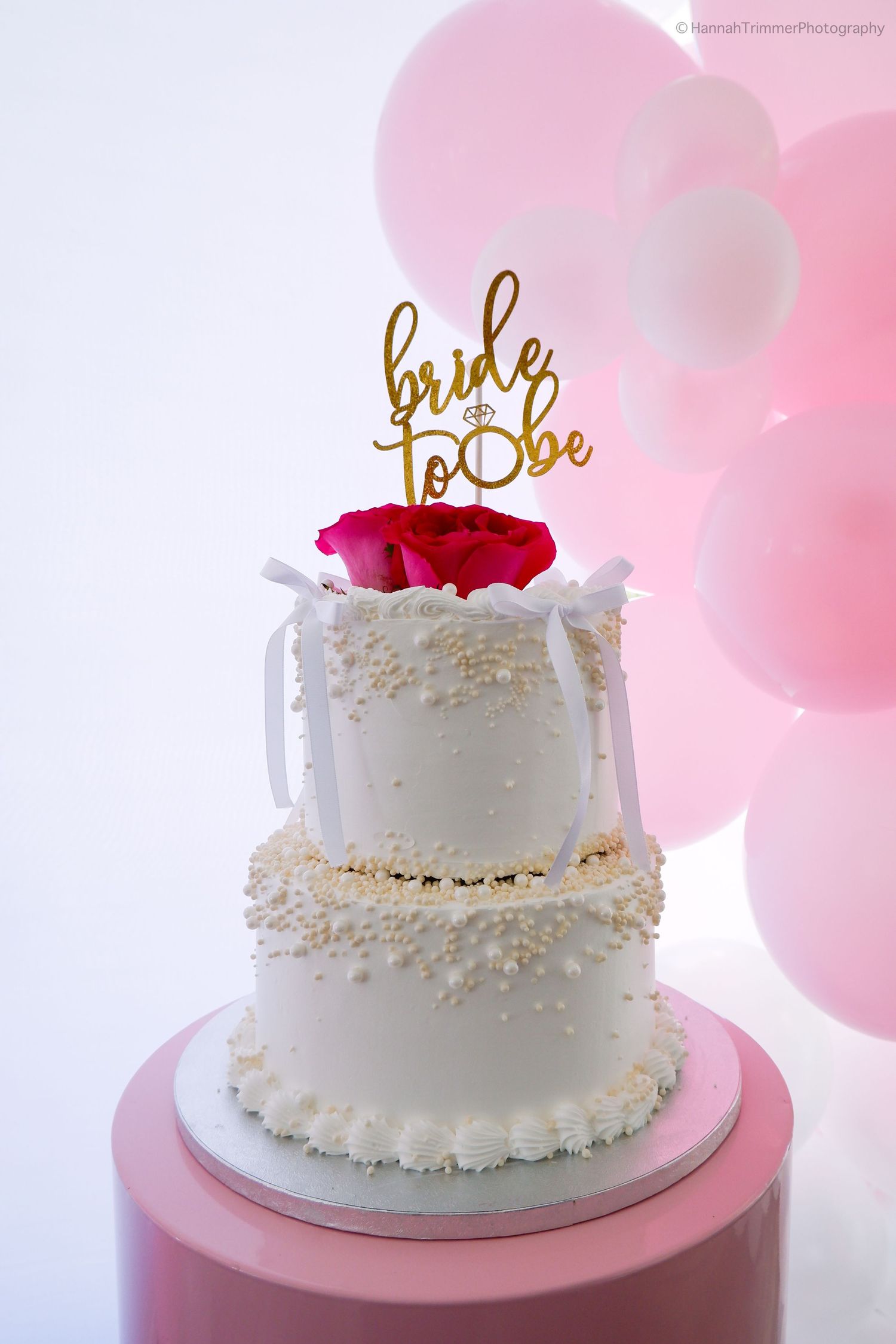 Two-tier white bridal shower cake with gold bride-to-be topper and pink rose.