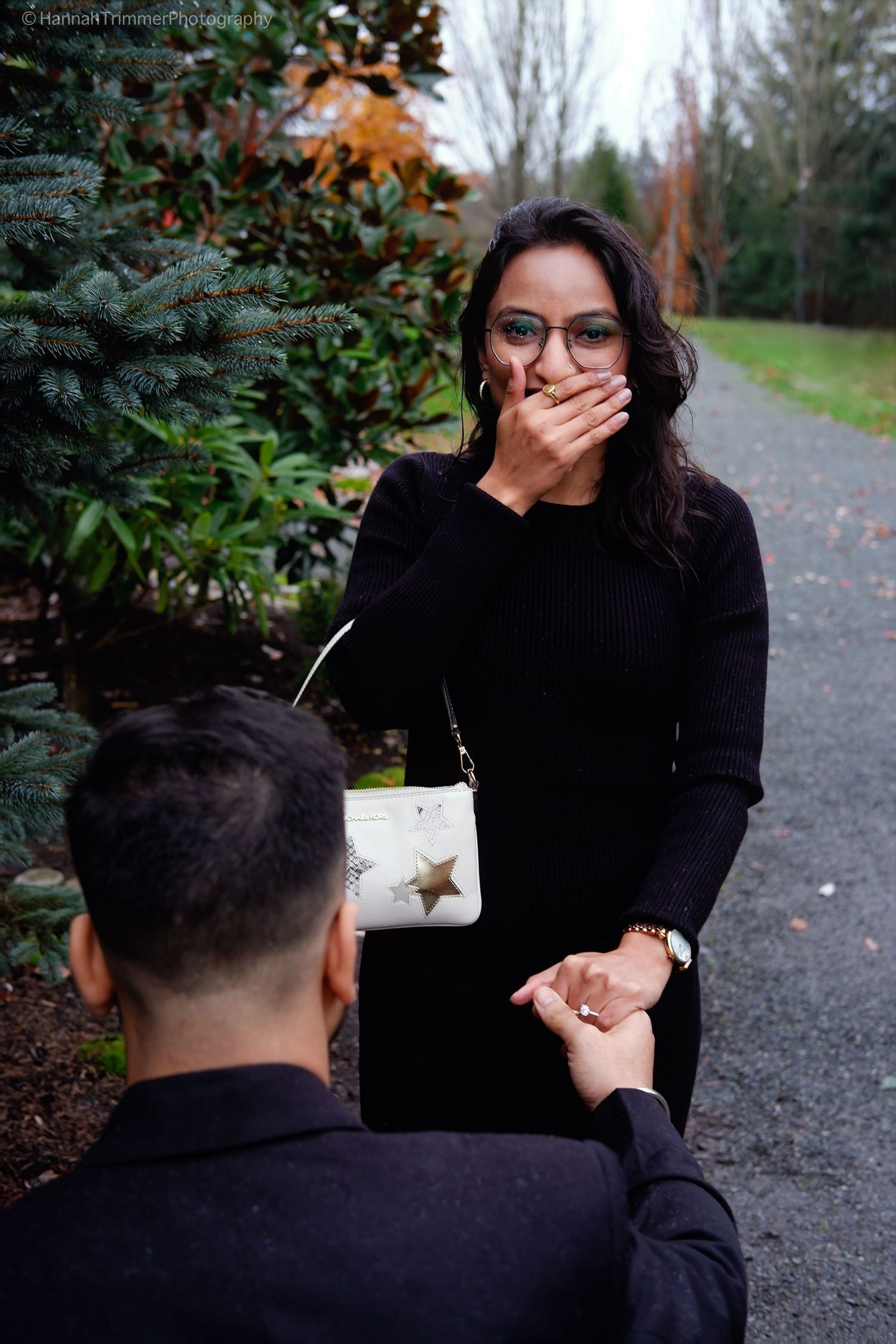Man proposing on one knee to shocked woman covering her mouth on a garden path.