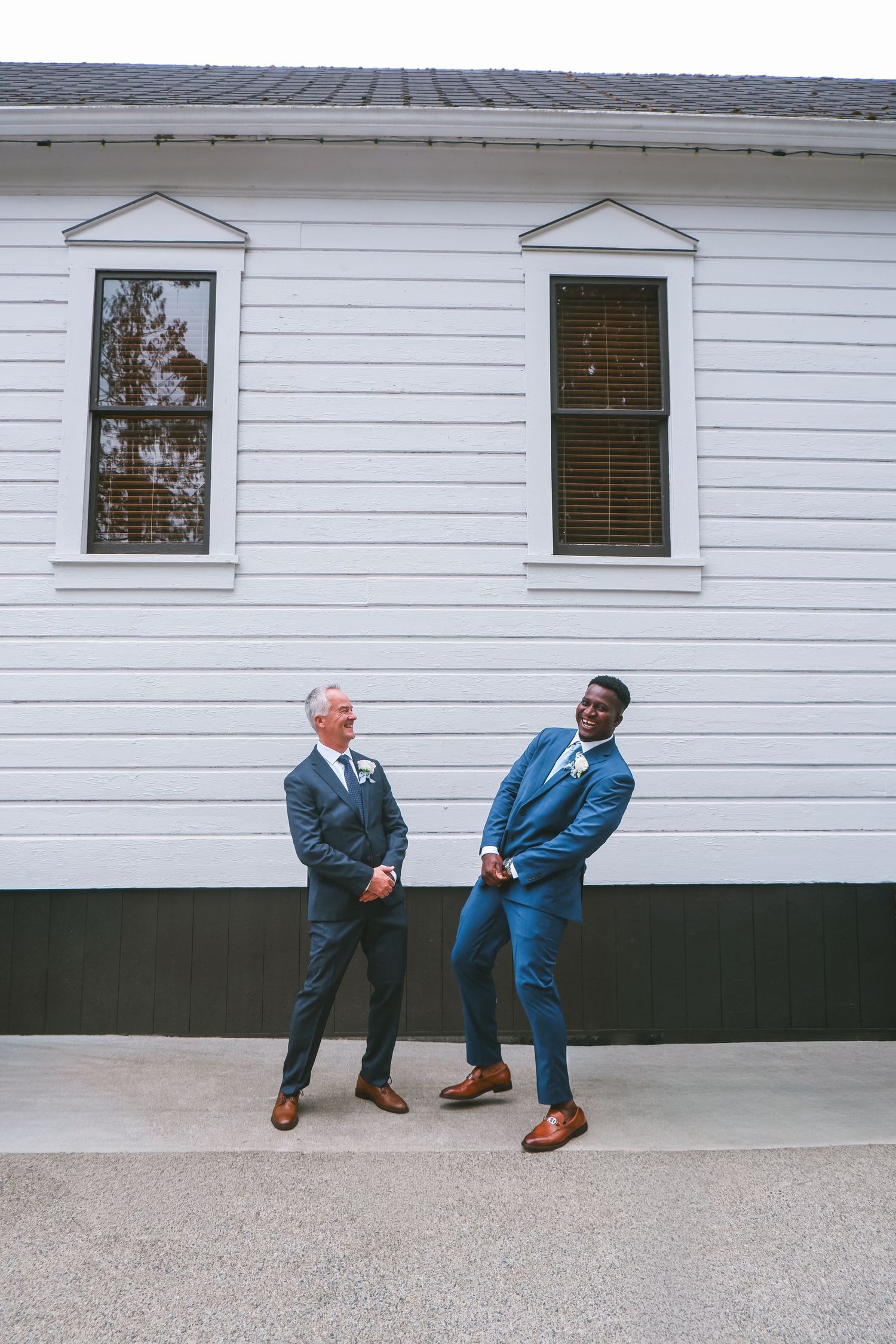 Two grooms in navy suits with brown shoes stand outside a white building on their wedding day.