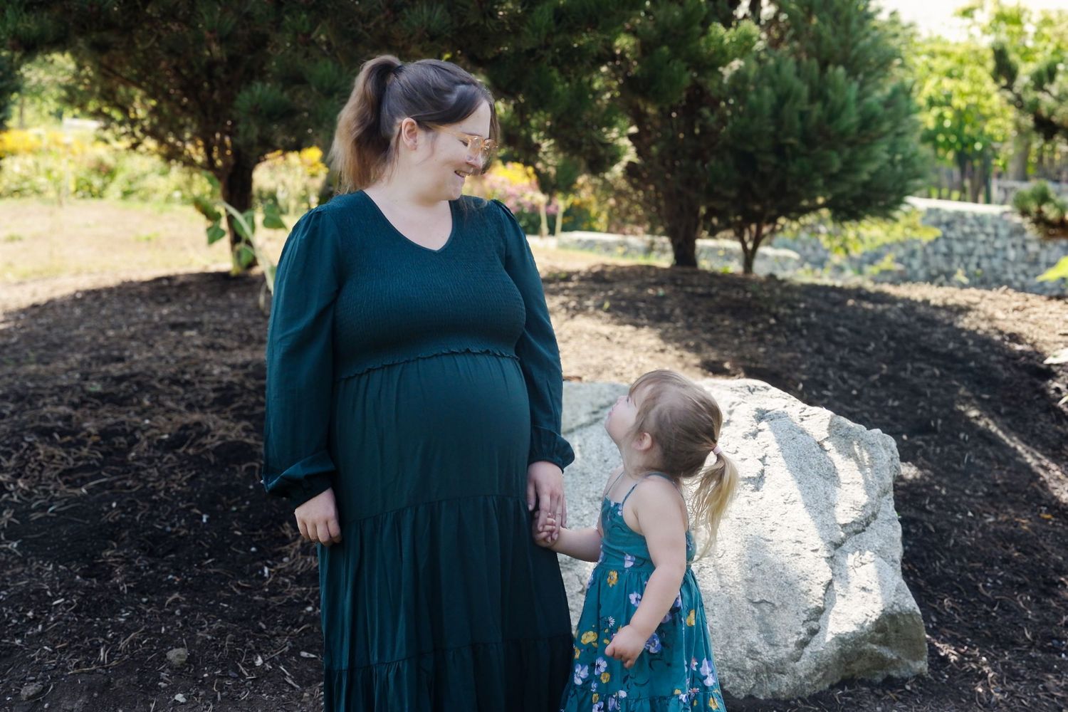 Pregnant woman in green dress smiling at toddler girl in floral dress outdoors near trees and large rock.