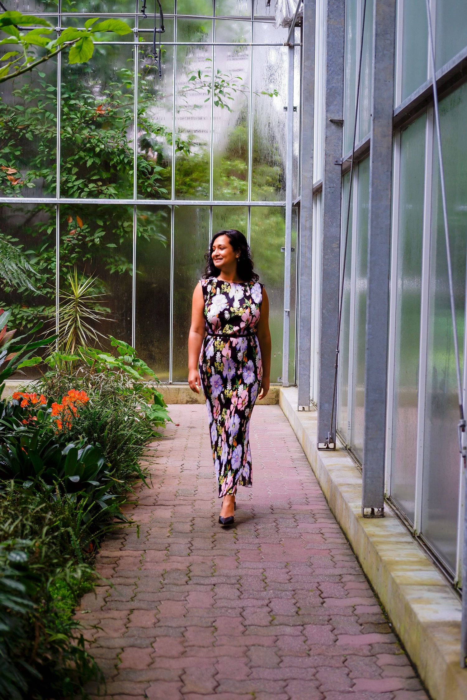 Woman in floral maxi dress walking through a lush greenhouse botanical garden pathway.