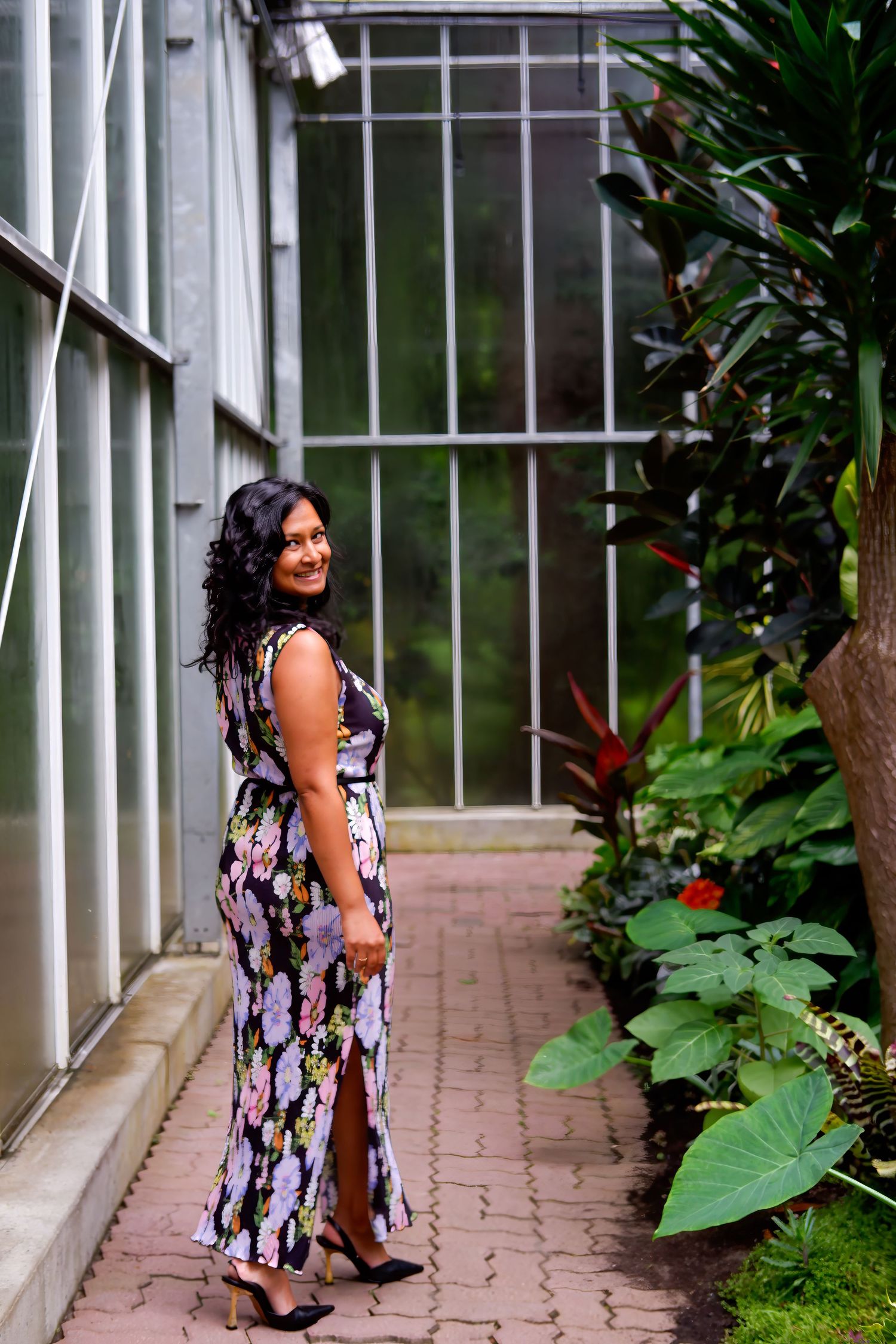 Woman in floral maxi dress smiling over shoulder inside a lush greenhouse garden.