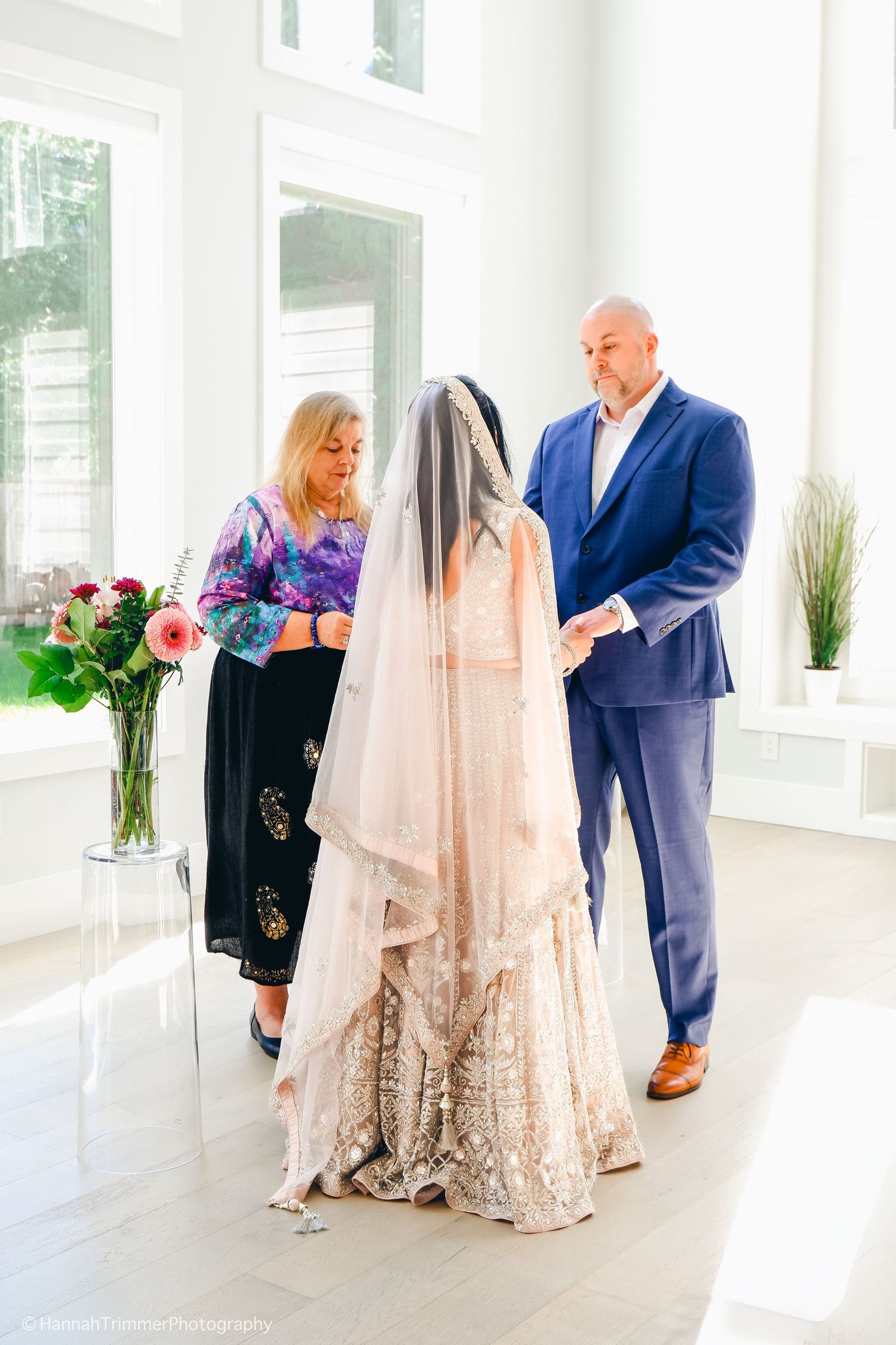 Officiant and groom adjusting bride's embroidered lehenga veil during elegant indoor wedding ceremony.