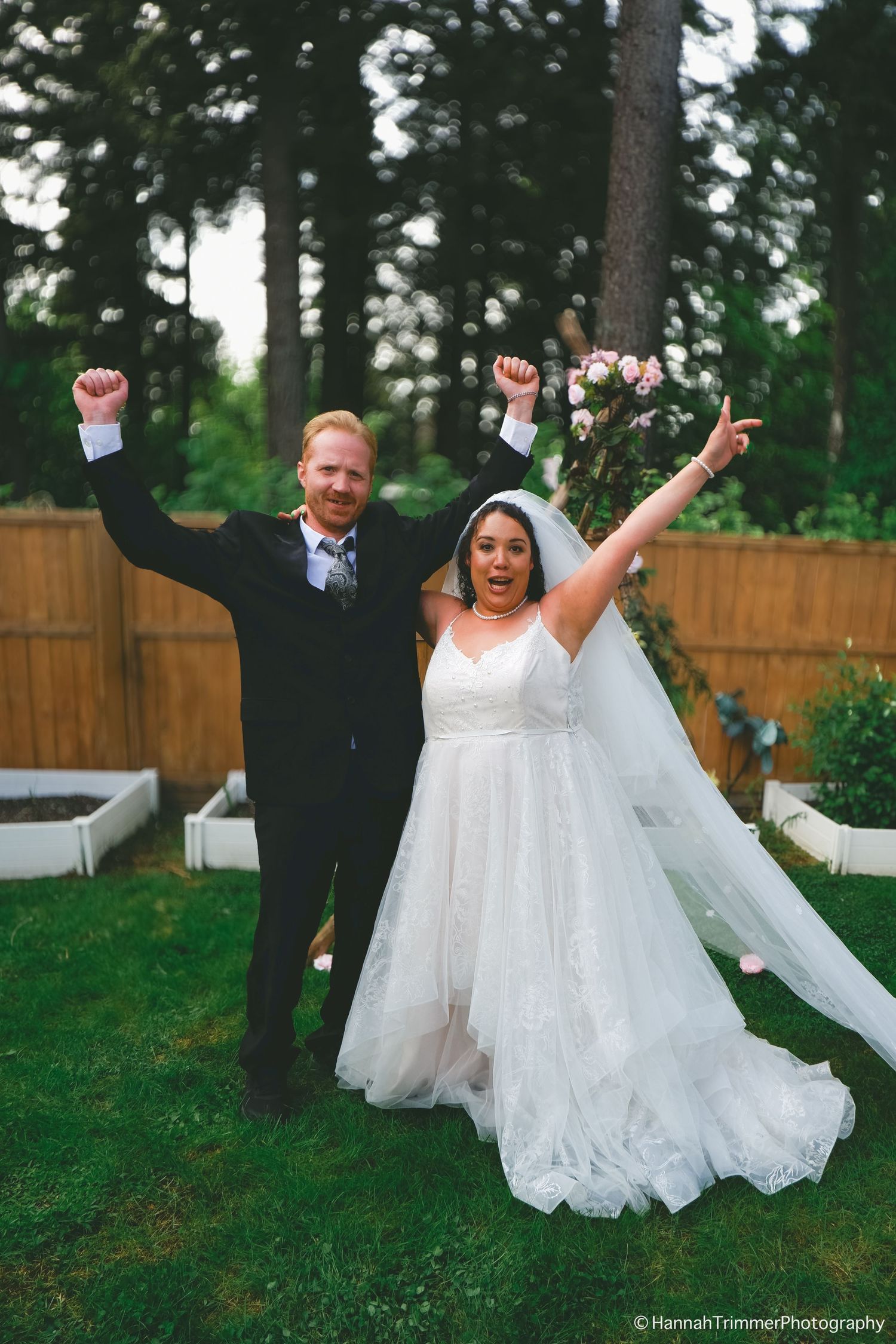 A joyful bride and groom celebrate with raised arms in a garden after their wedding ceremony.