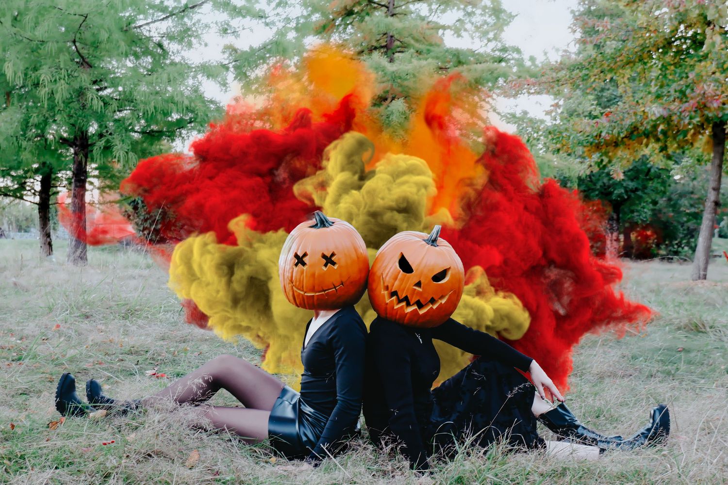Two people wearing jack-o-lantern heads sit back-to-back outdoors surrounded by red and yellow Halloween smoke.