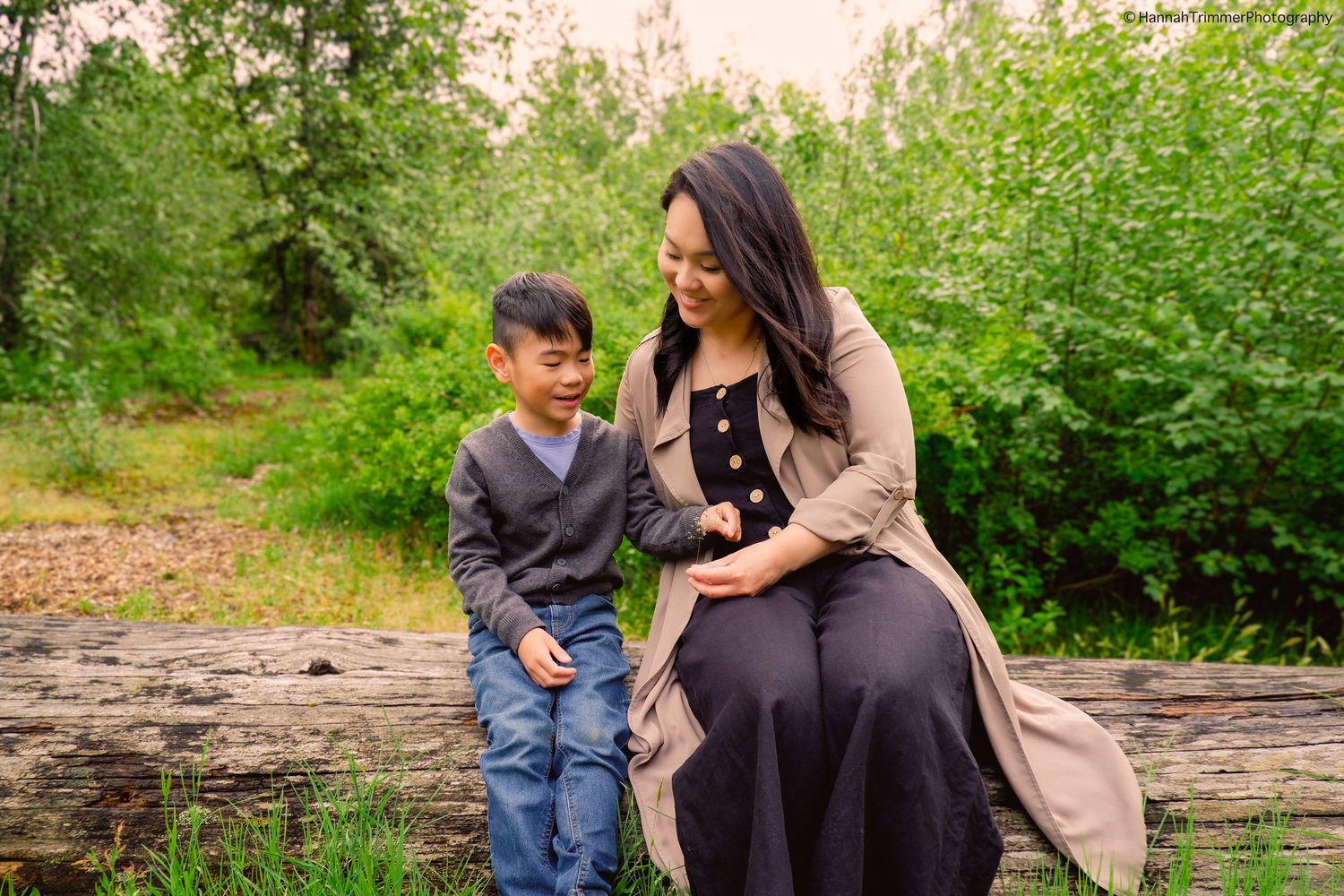 Mother and young son sharing a sweet moment seated on a log in a green outdoor setting.