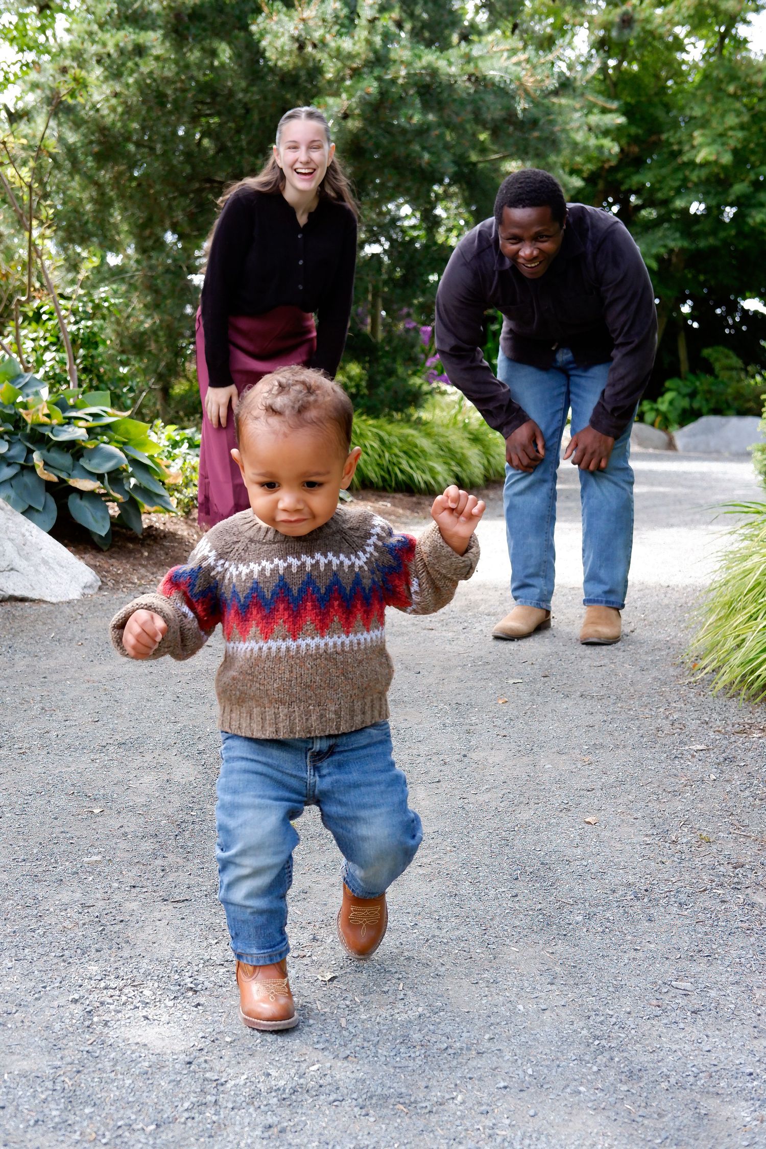 Joyful interracial parents encouraging their toddler to walk toward them on a garden path.
