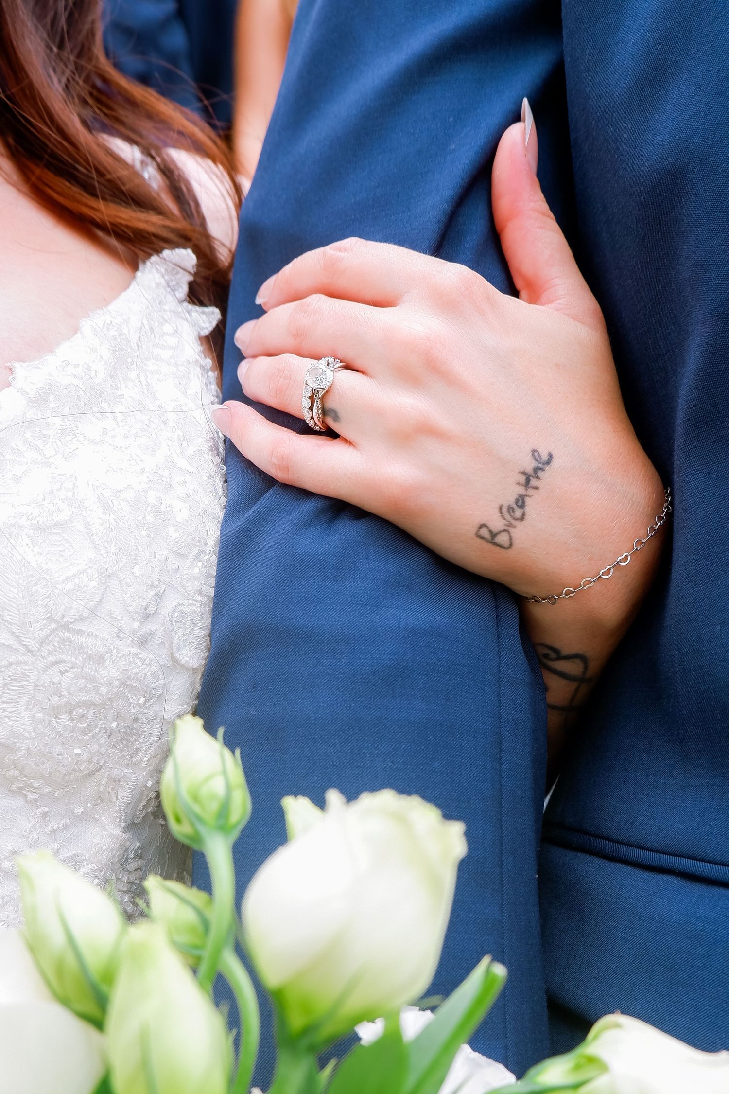 Bride showing diamond engagement ring and wrist tattoo while holding groom's arm in blue suit at wedding.