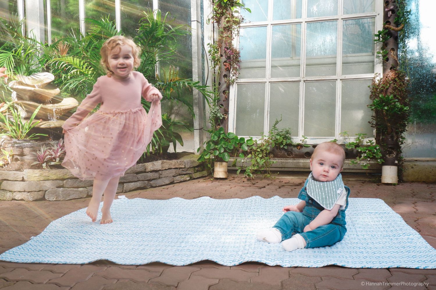 Two young children, a toddler girl in pink tutu and baby boy, play on blue mat in greenhouse.