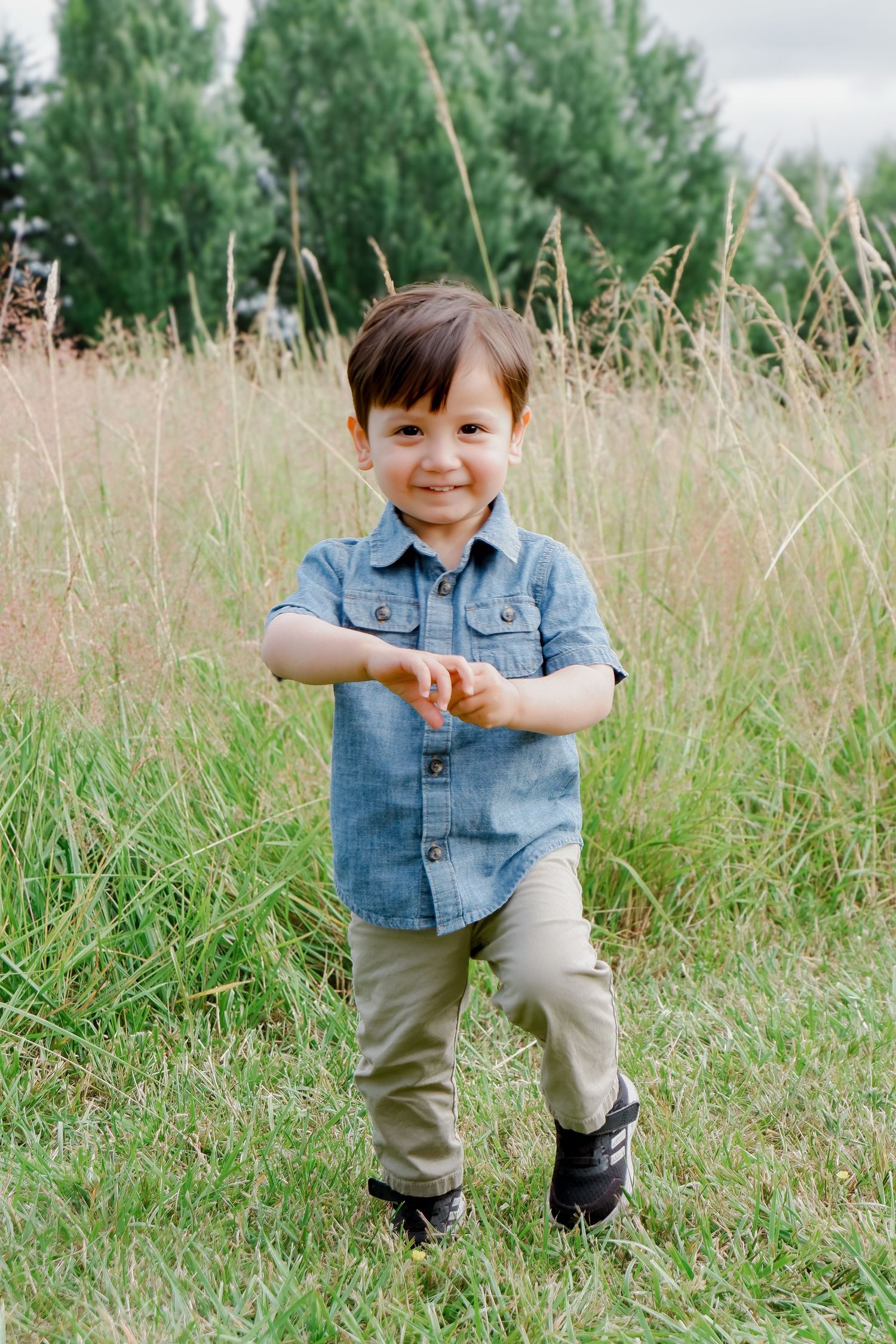 Smiling toddler boy in a blue denim shirt standing in a tall grass field outdoors.