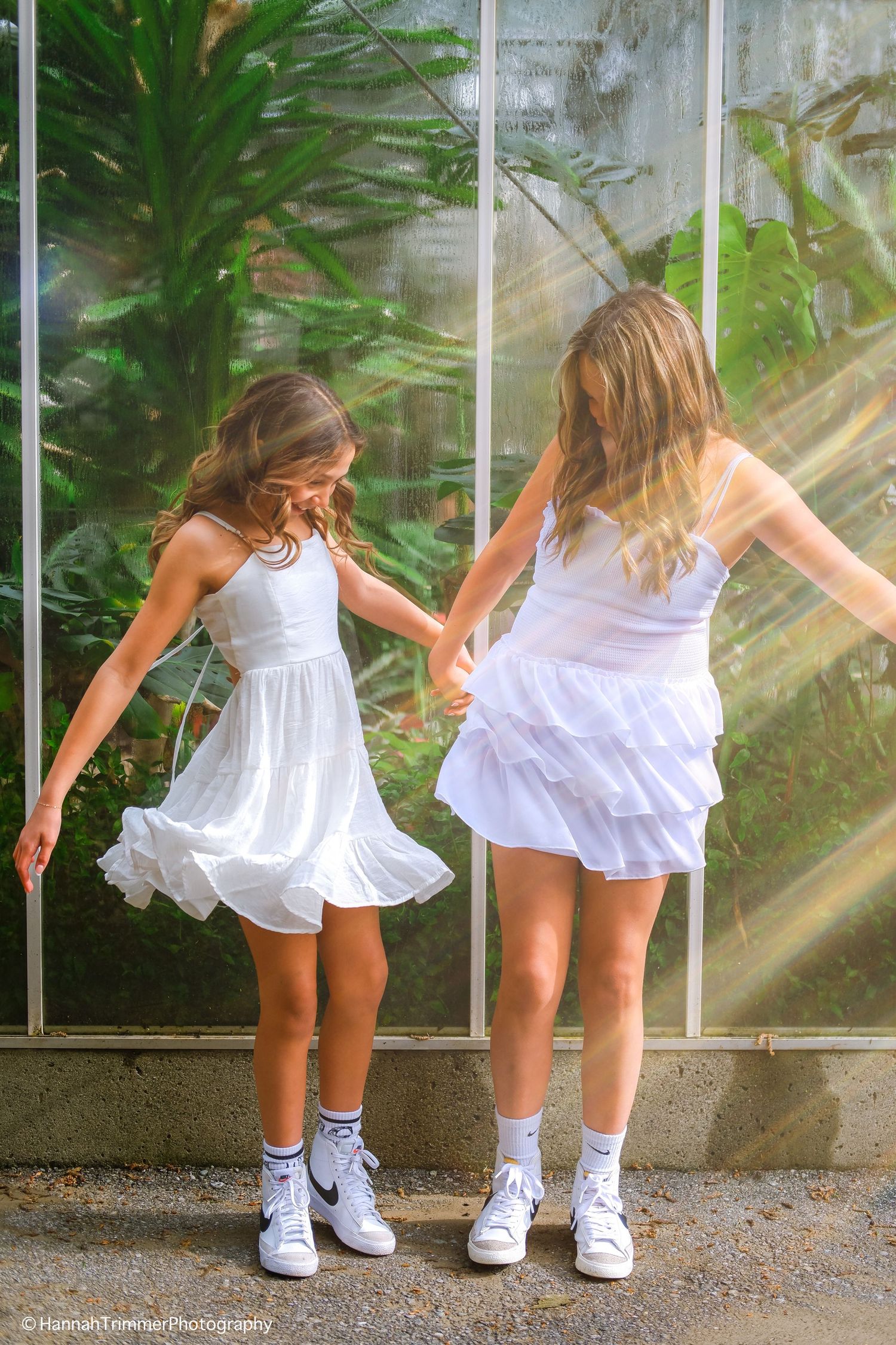 Two girls in white dresses and Nike sneakers hold hands dancing near a sunlit greenhouse glass wall.