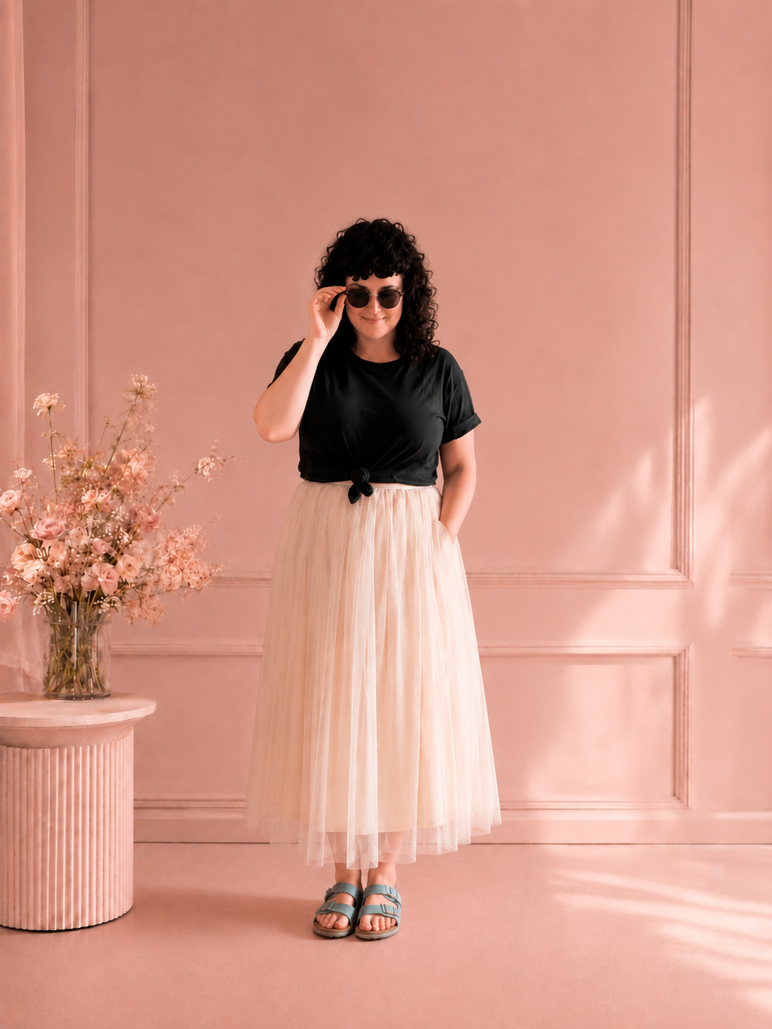 Woman in black tee and cream tulle skirt wearing sunglasses against a pink wall with flowers.