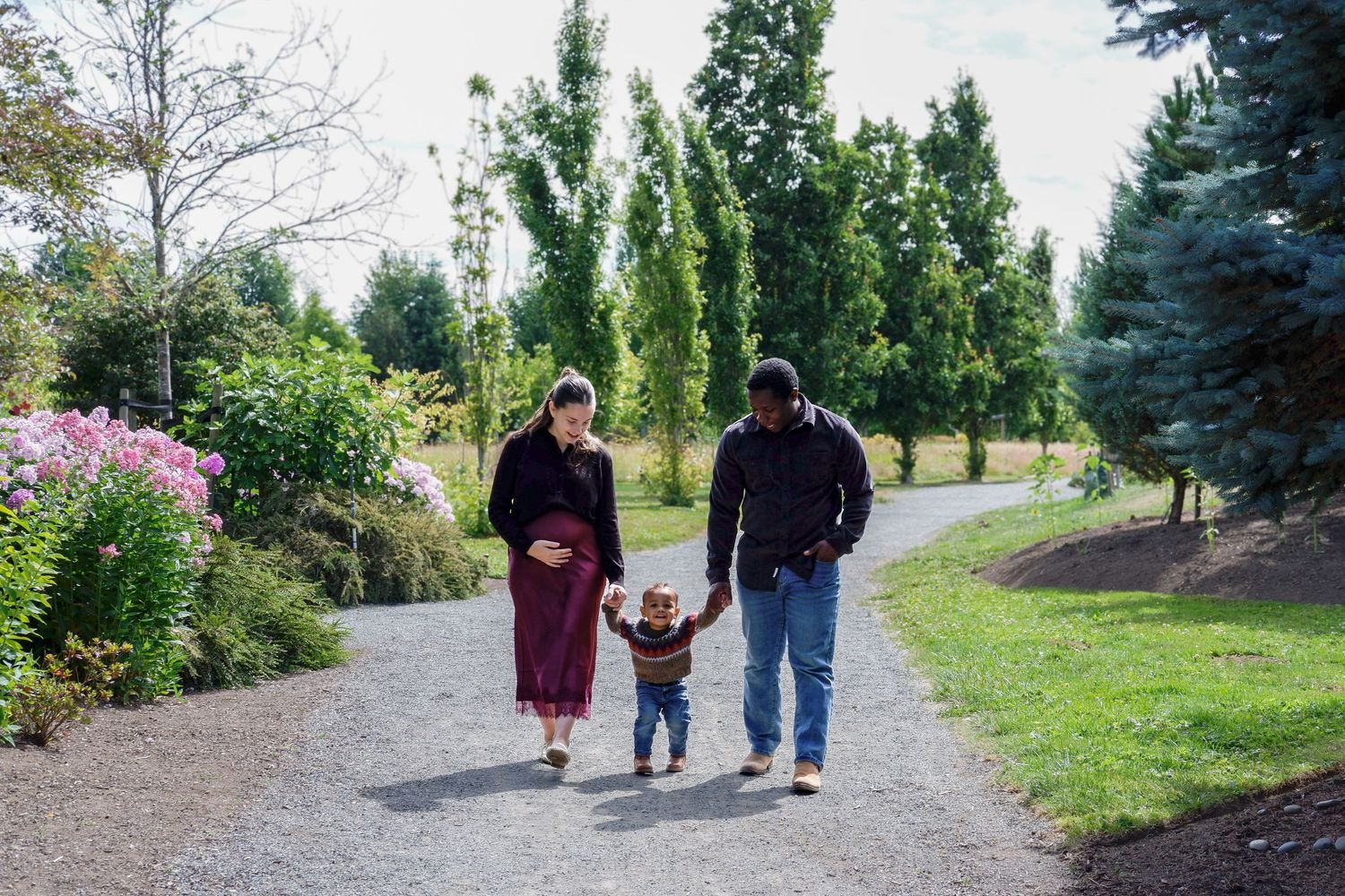 Expecting couple walking hand-in-hand with toddler on garden path surrounded by flowers.