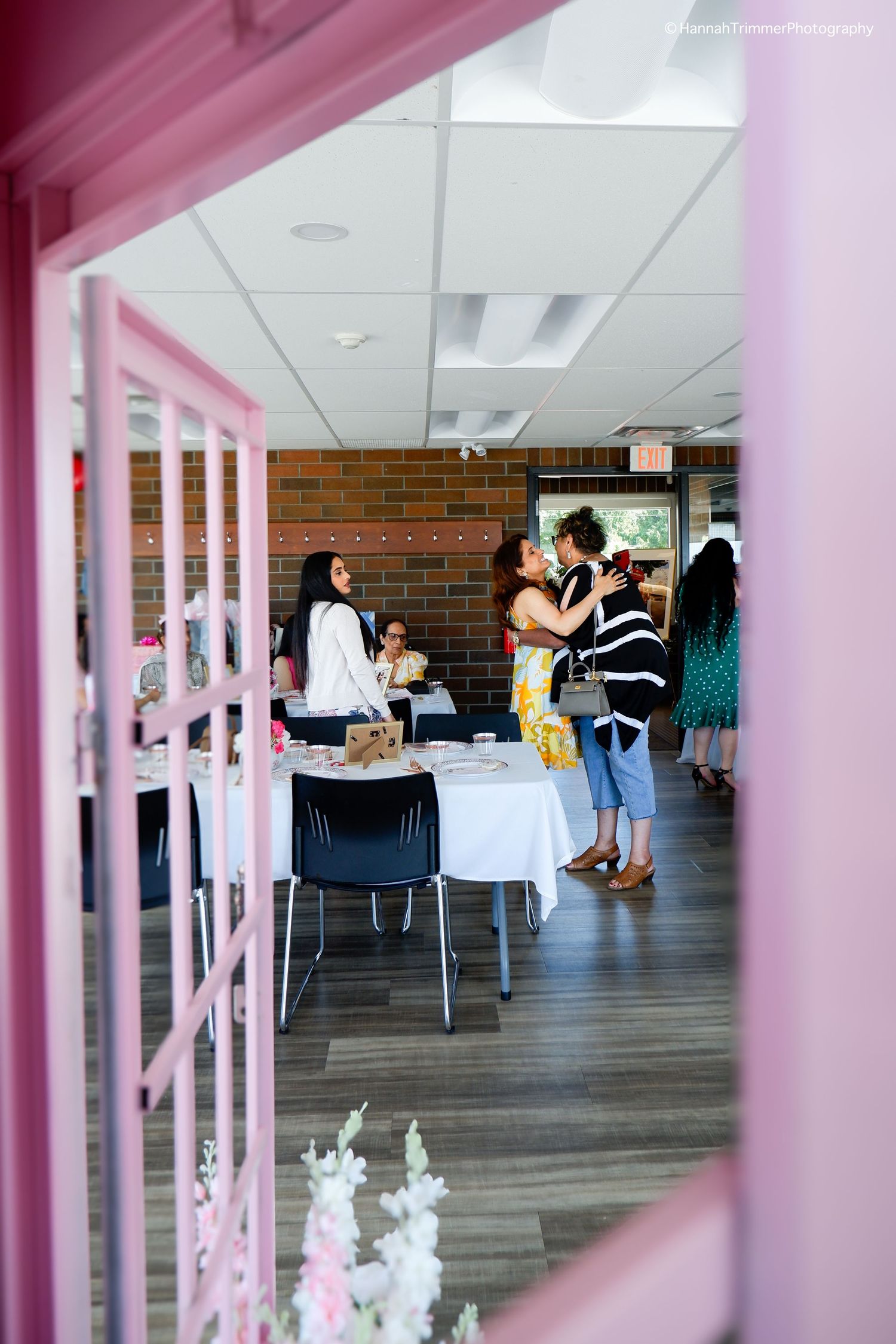 Guests mingle and embrace at a bridal shower event viewed through a pink window frame.