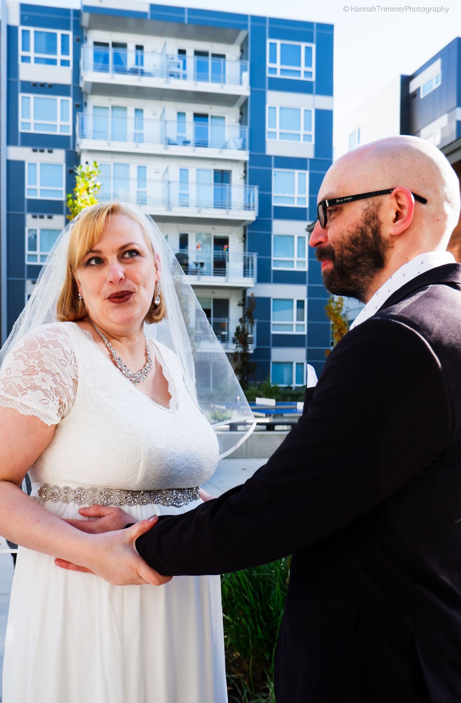 Bride in white lace wedding dress holds hands with bald groom in black suit outside modern apartment building.
