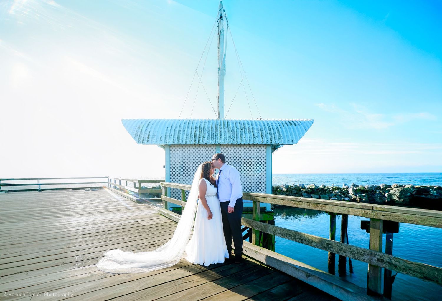 Bride and groom share a romantic kiss on a waterfront pier with a small boathouse and ocean view on their wedding day.