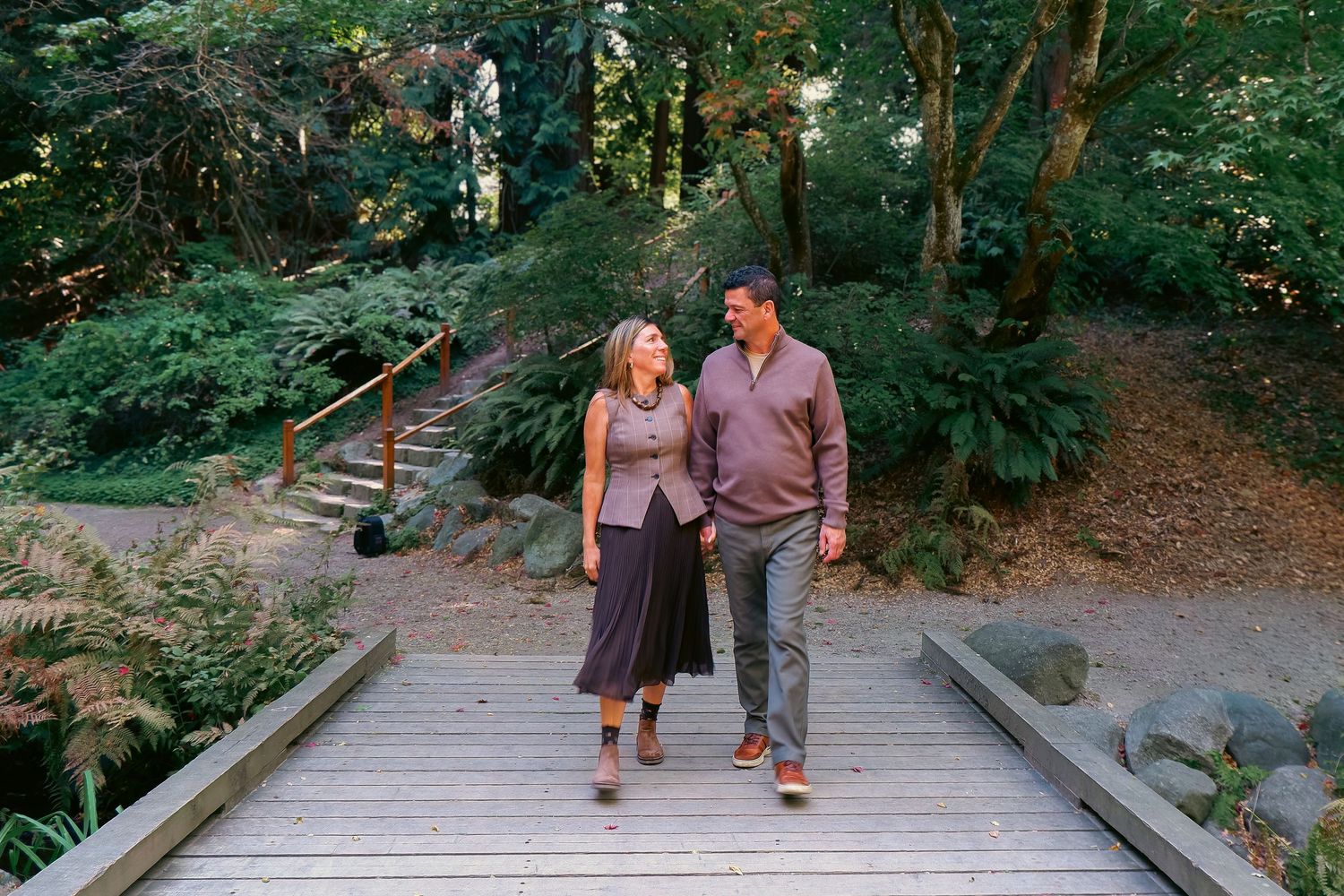 Couple holding hands and walking on a wooden bridge path surrounded by lush green trees and ferns.