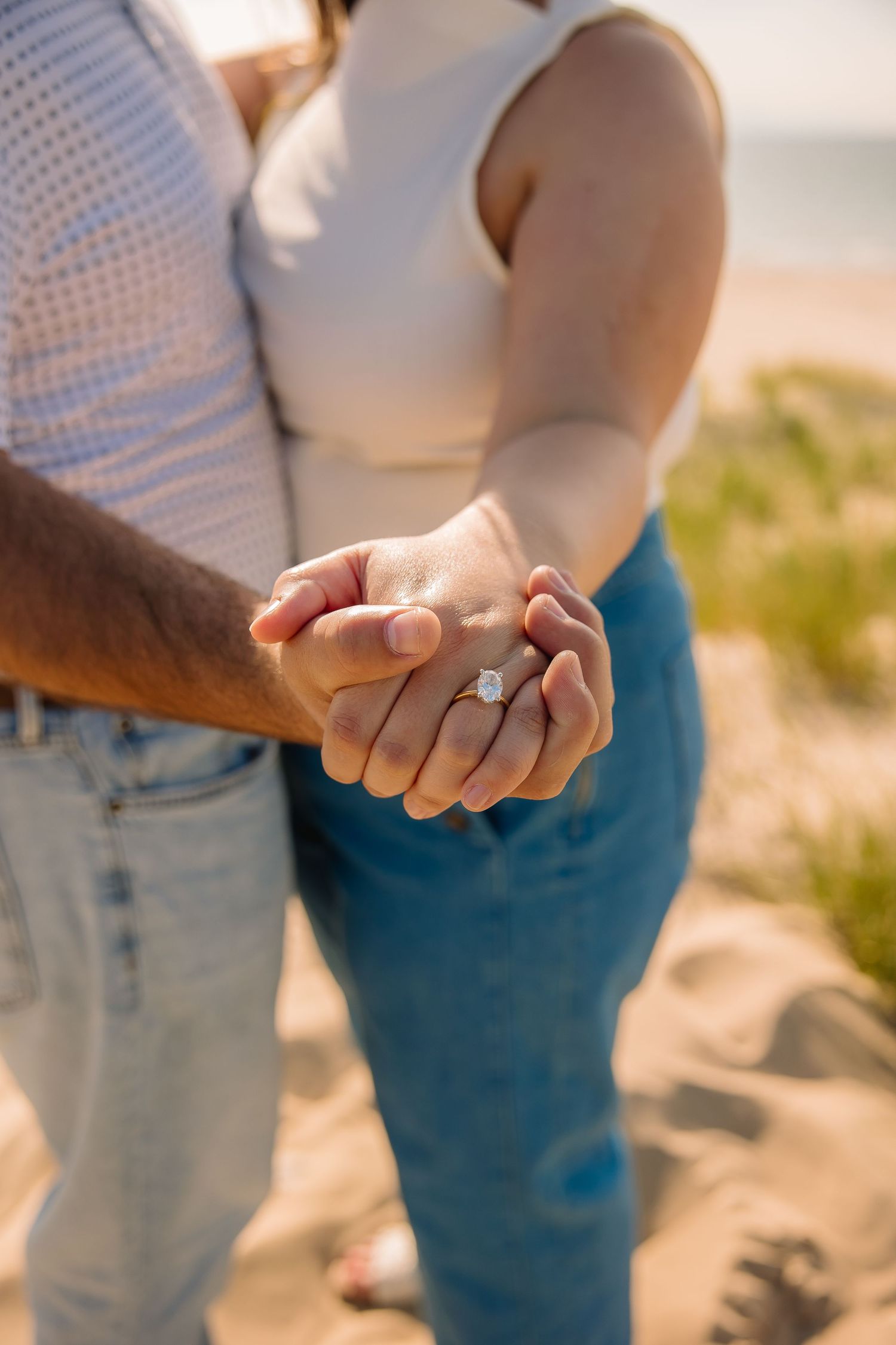 Couple holding hands on a sandy beach displaying an engagement ring, wearing jeans and casual tops.