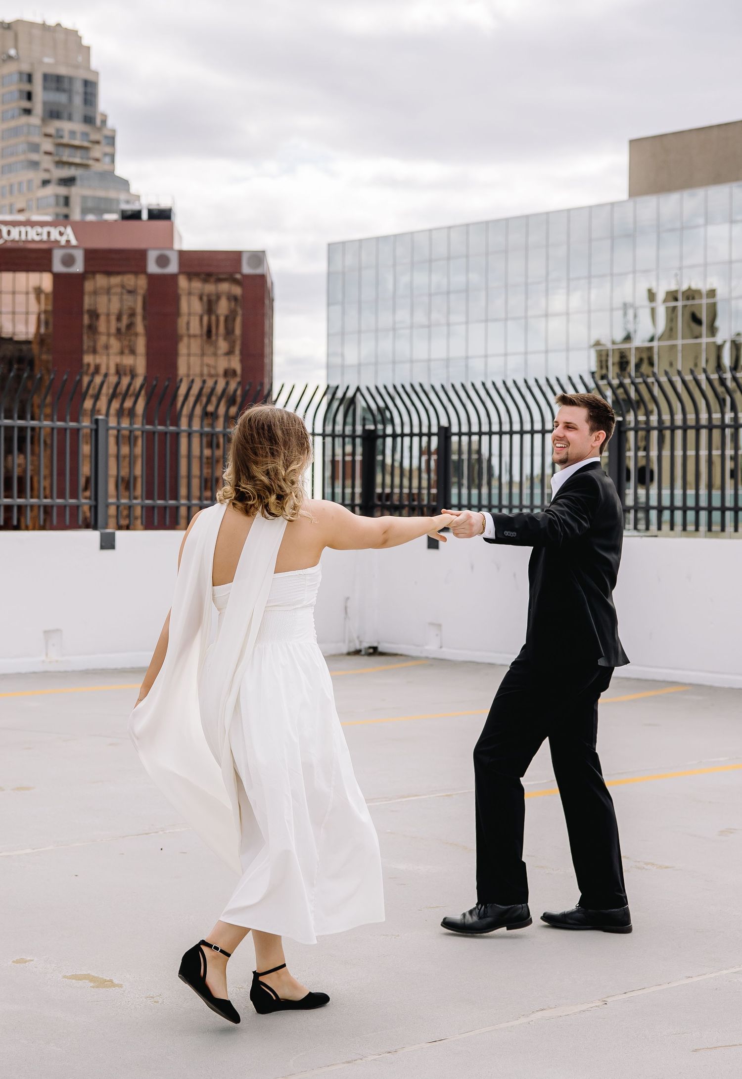 Bride and groom dancing on rooftop with city skyline backdrop, bride in white dress with cape, groom in black tuxedo.