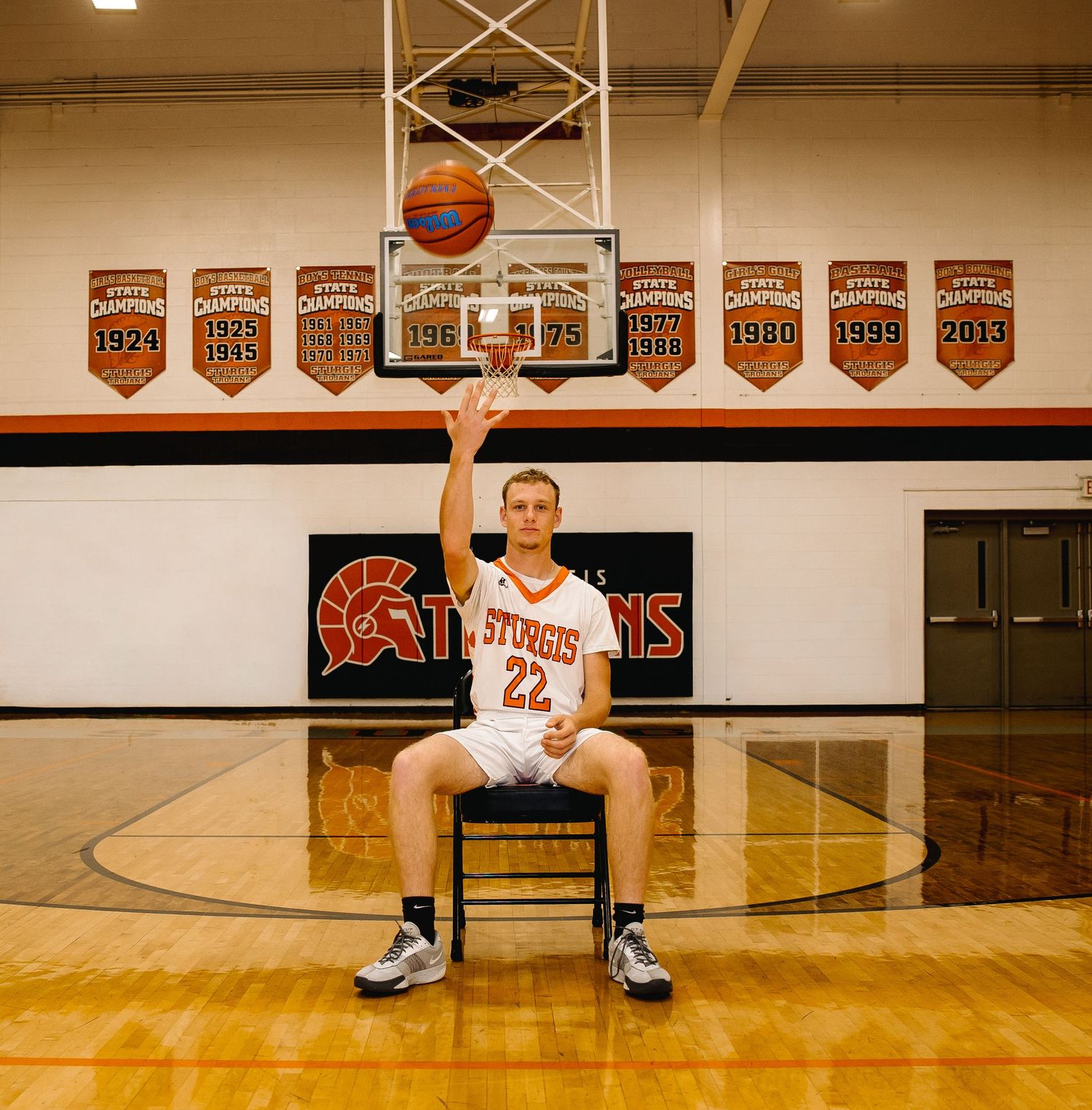 Basketball player #22 sits on a chair under the hoop in a gym with championship banners for the Sturgis team.