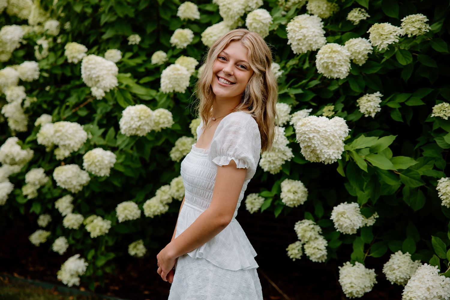 Smiling blonde woman in white dress poses before lush hydrangea bushes in bloom.