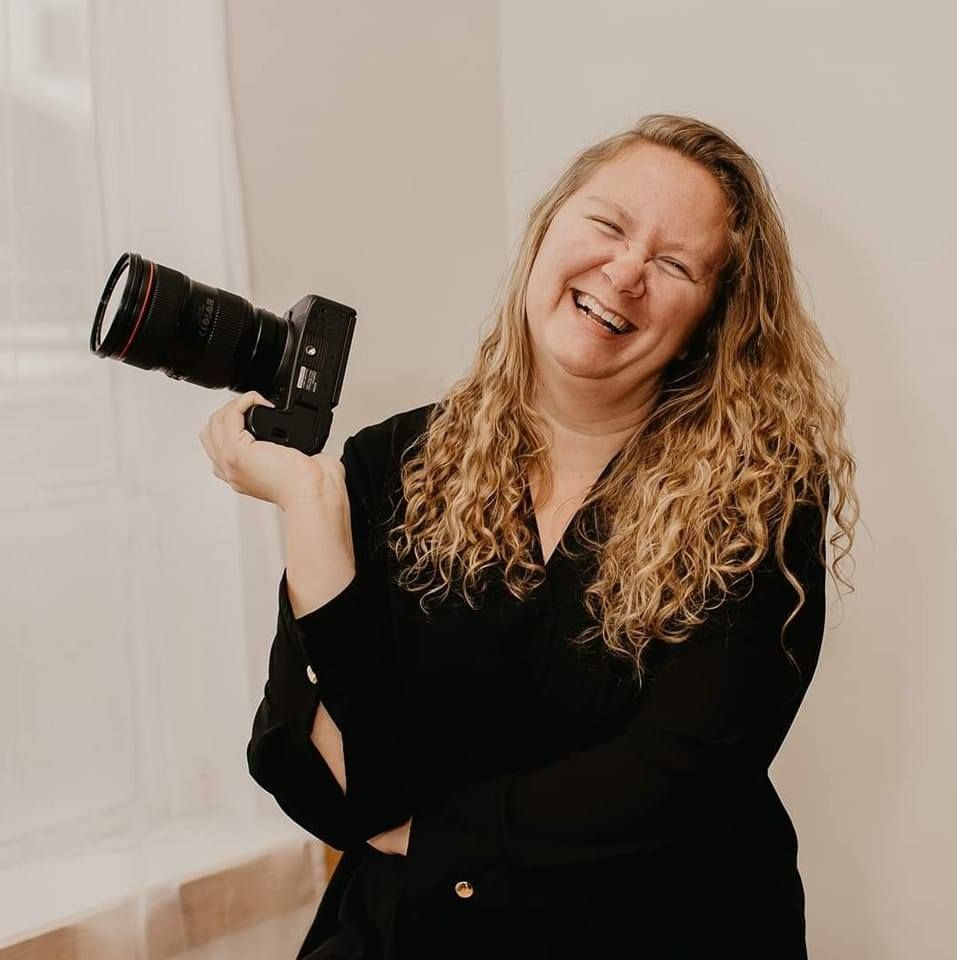 Smiling woman with curly blonde hair holding a Canon DSLR camera, wearing a black top in a bright studio.