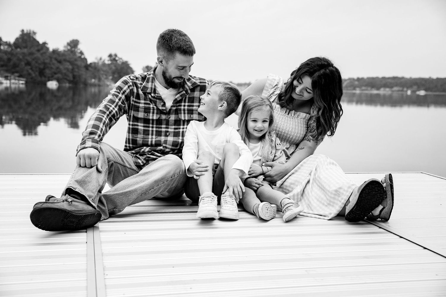 Black and white photo of a happy family sitting on a dock by a lake, parents and two young children smiling together.