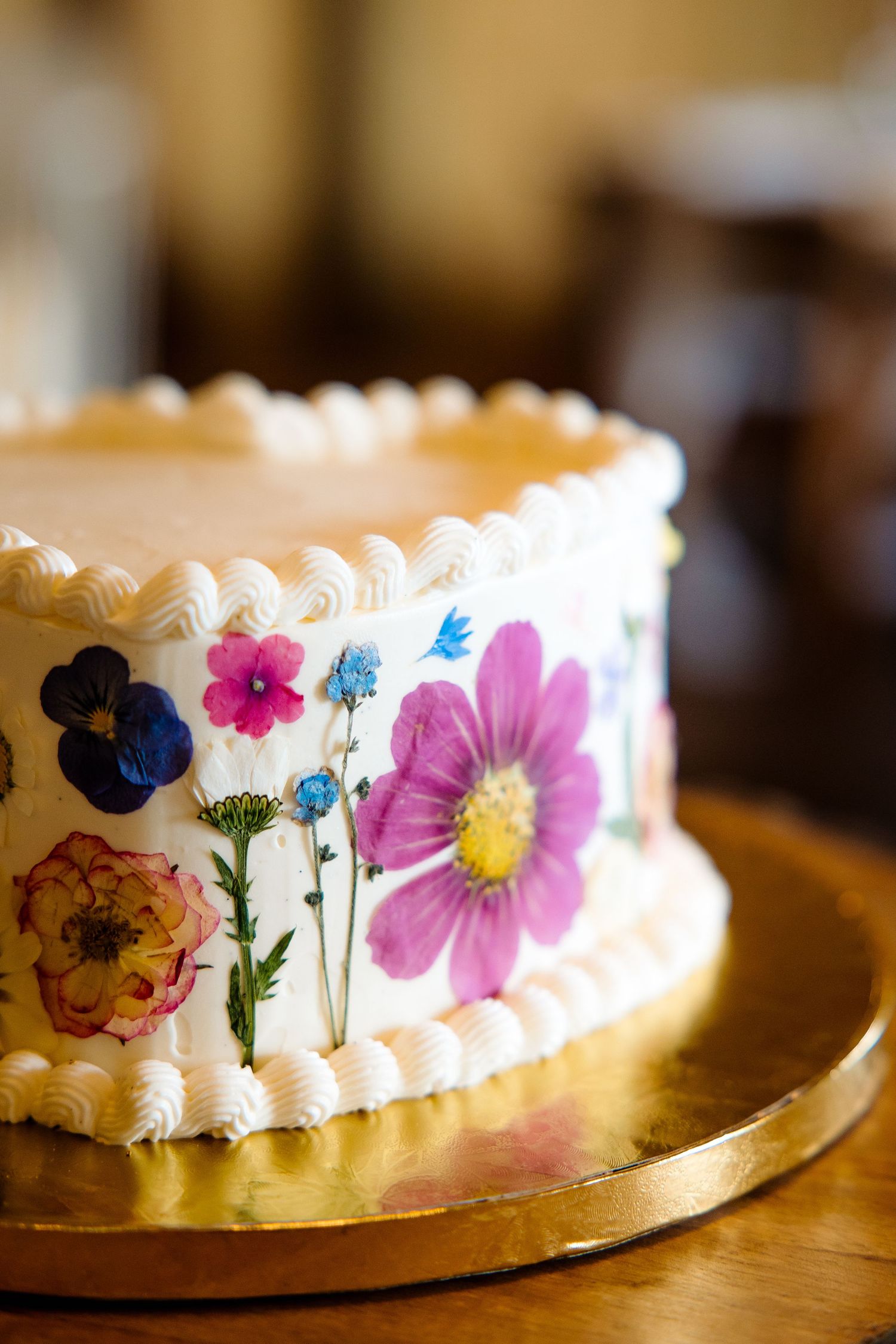 White frosted cake decorated with colorful pressed flowers including pink cosmos, blue pansy, and wildflowers on a gold cake board.