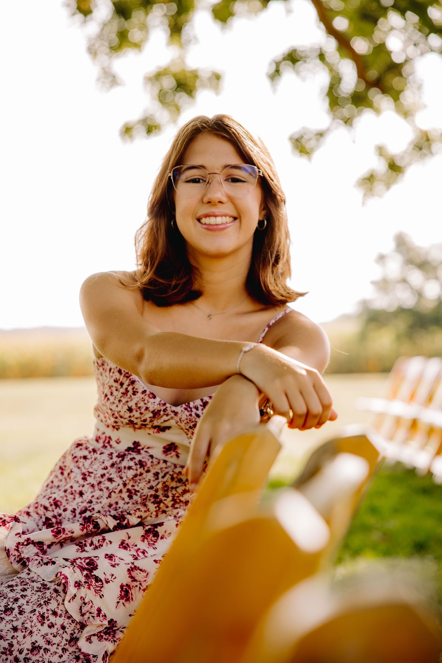 Smiling young woman in floral dress and glasses sitting outdoors on a sunny day near a bench under a tree.